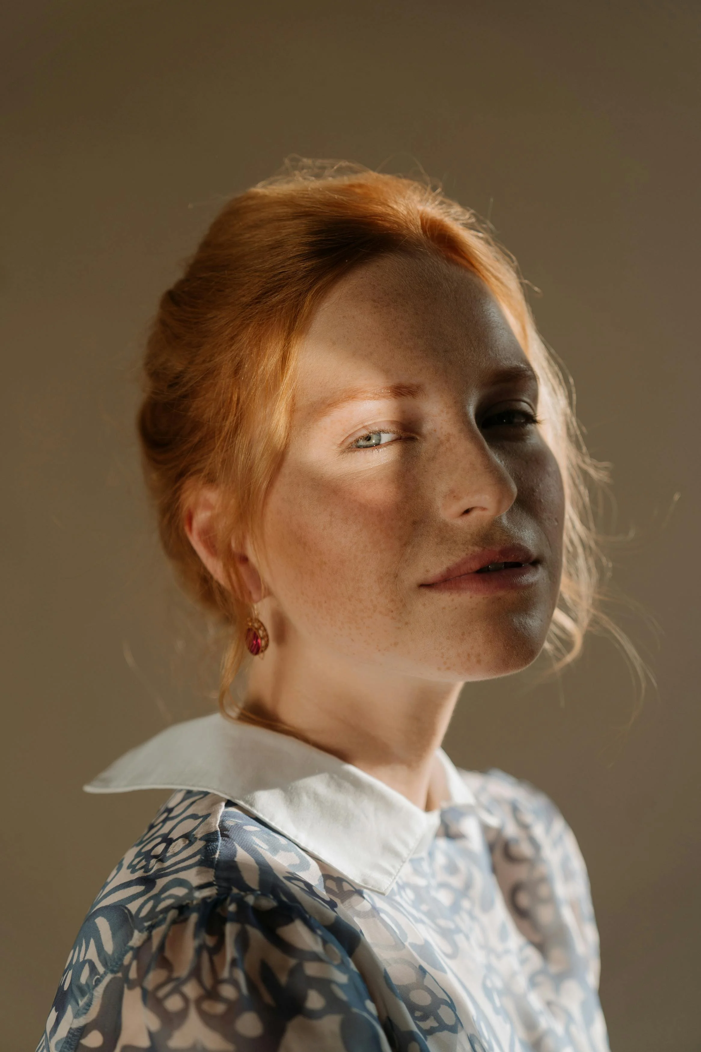 Close-up portrait of a woman with red hair and freckles, wearing a patterned shirt and white collar, with sunlight casting shadows on her face.
