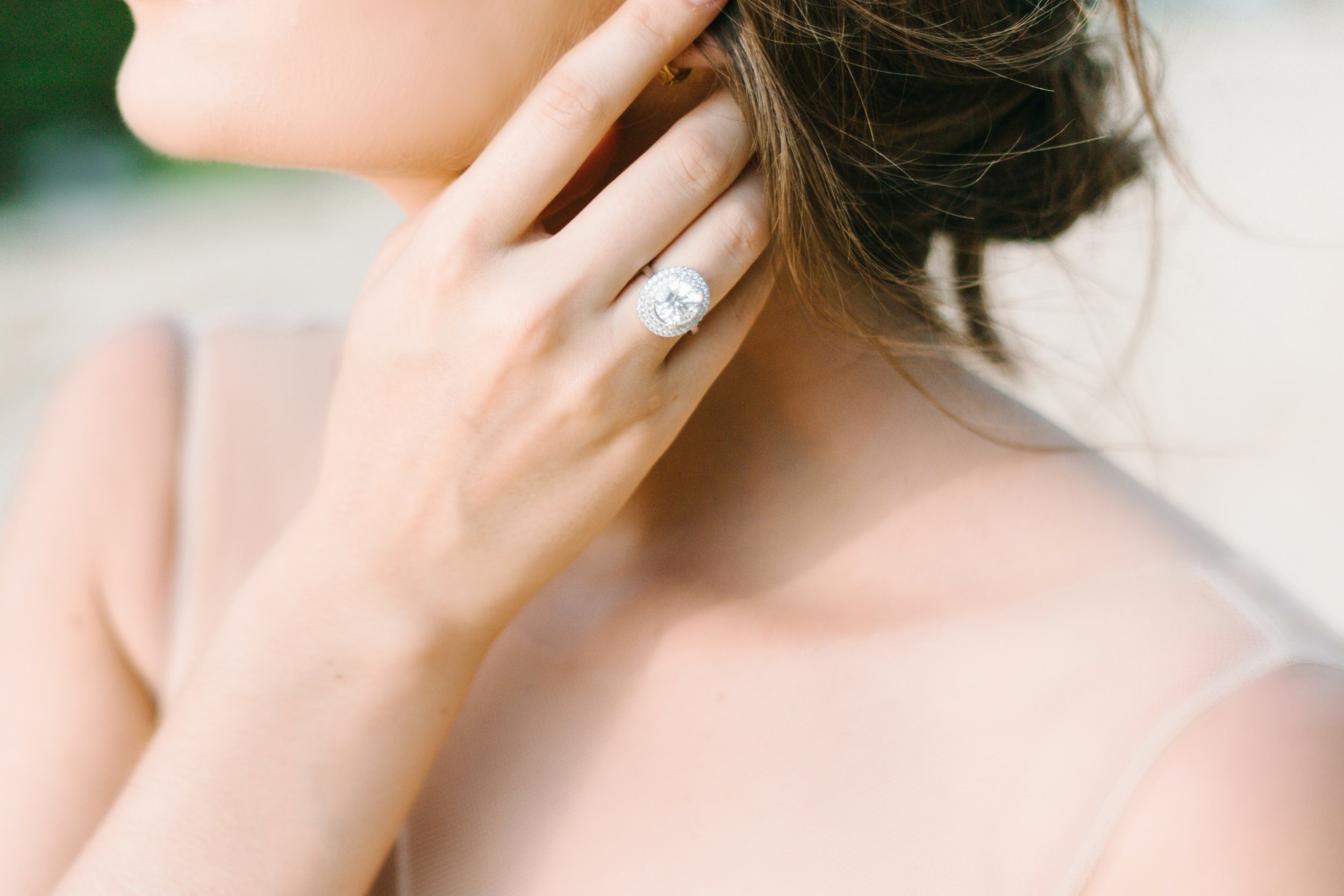 Close-up of a woman's face and hand, showing a large, sparkling diamond ring on her finger, with a blurred background.