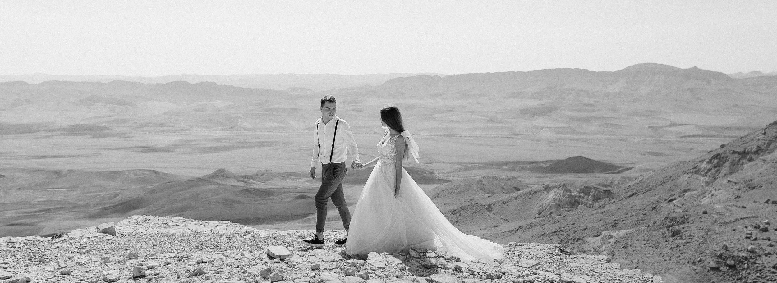 A couple in wedding attire holding hands on a rocky hilltop in a barren, mountainous landscape.