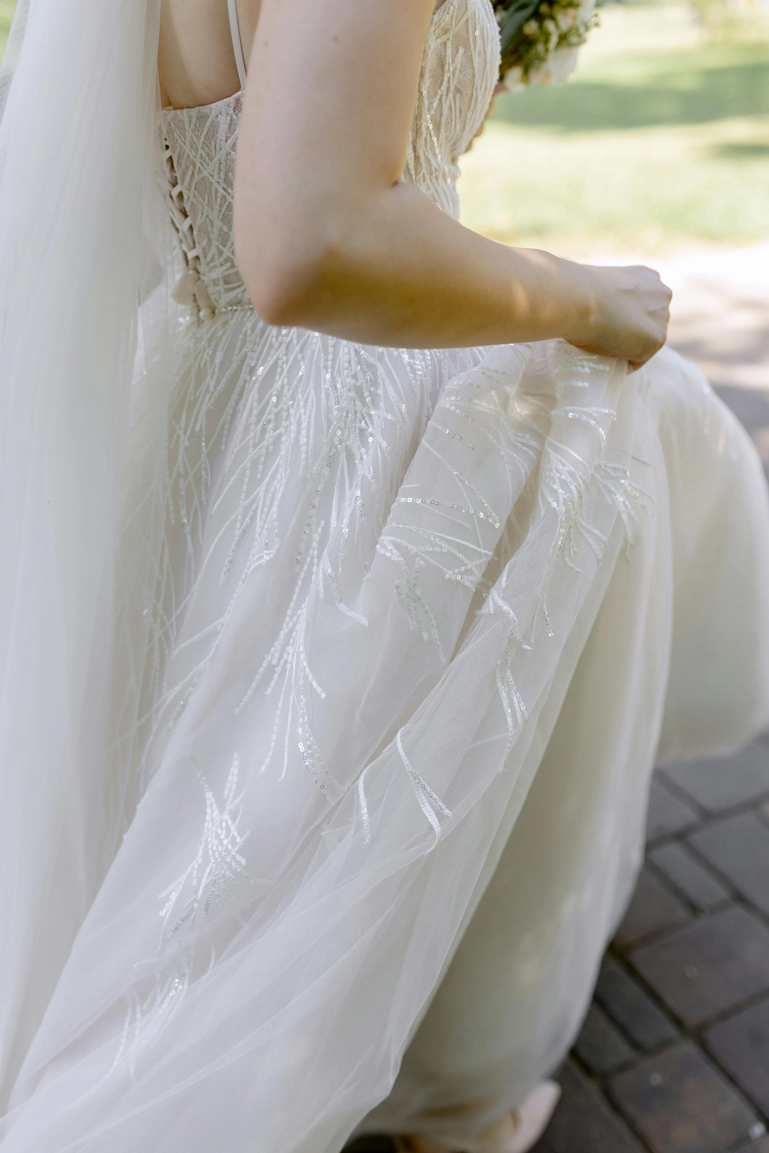 Close-up of a bride holding her wedding dress, which features intricate embroidery and sequins, outdoors on a sunny day.