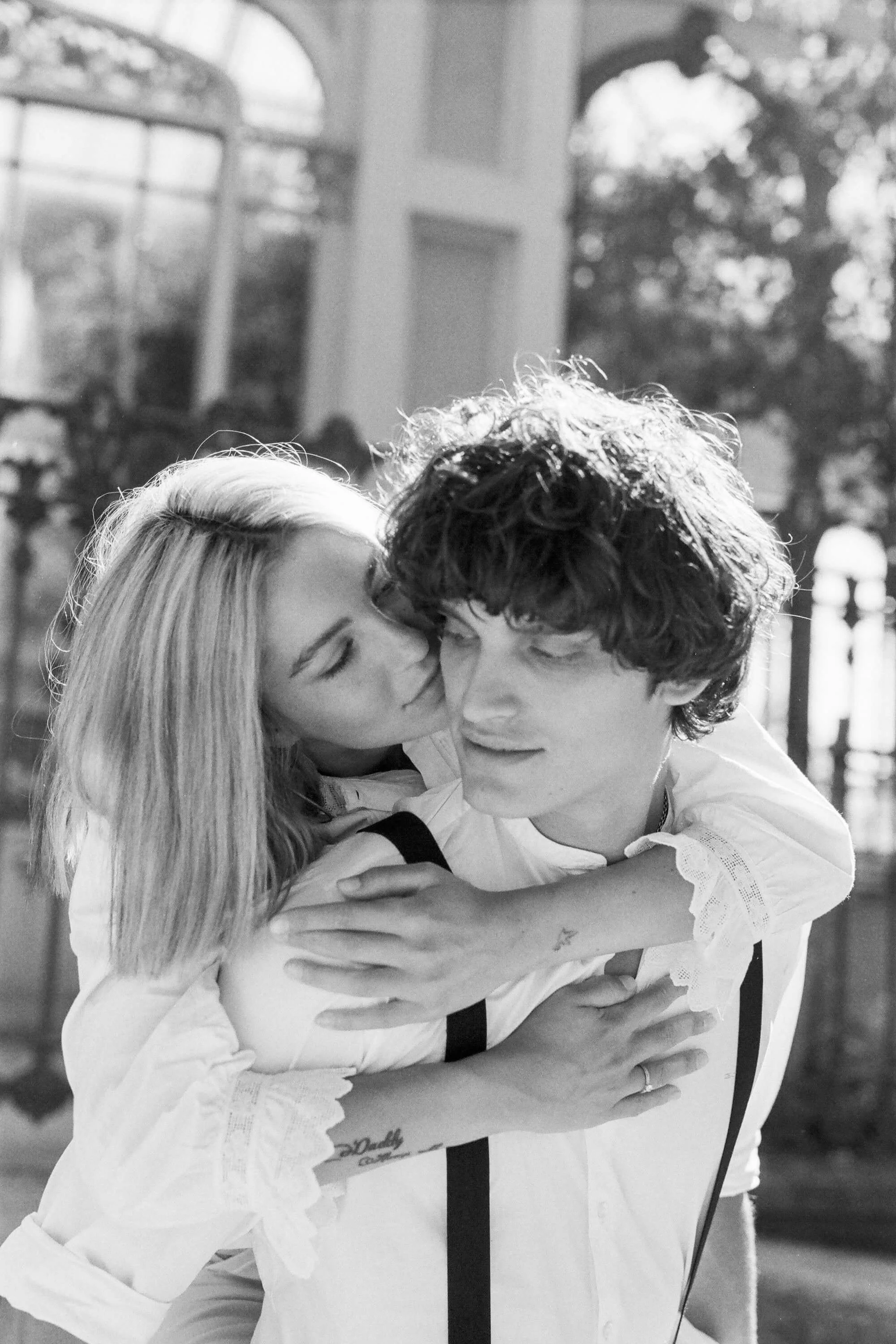 A black-and-white photo of a couple, with the woman kissing the man on the cheek, both smiling and embracing outdoors.