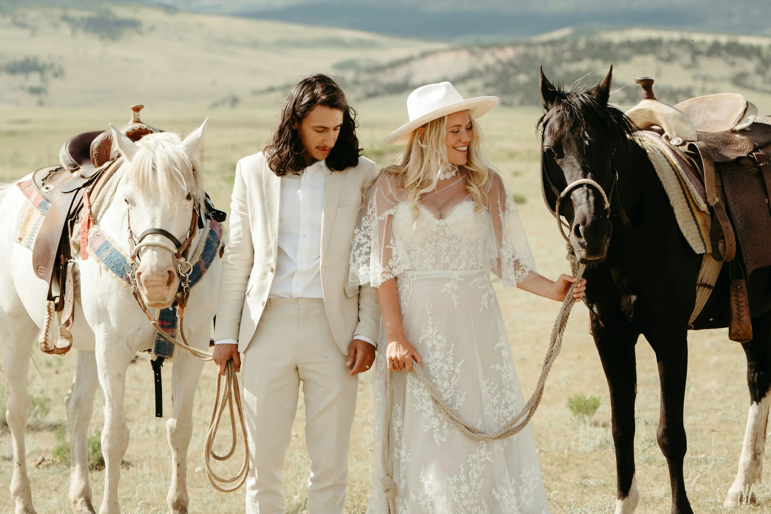 A couple in wedding attire standing outdoors with two horses on a grassy plain.