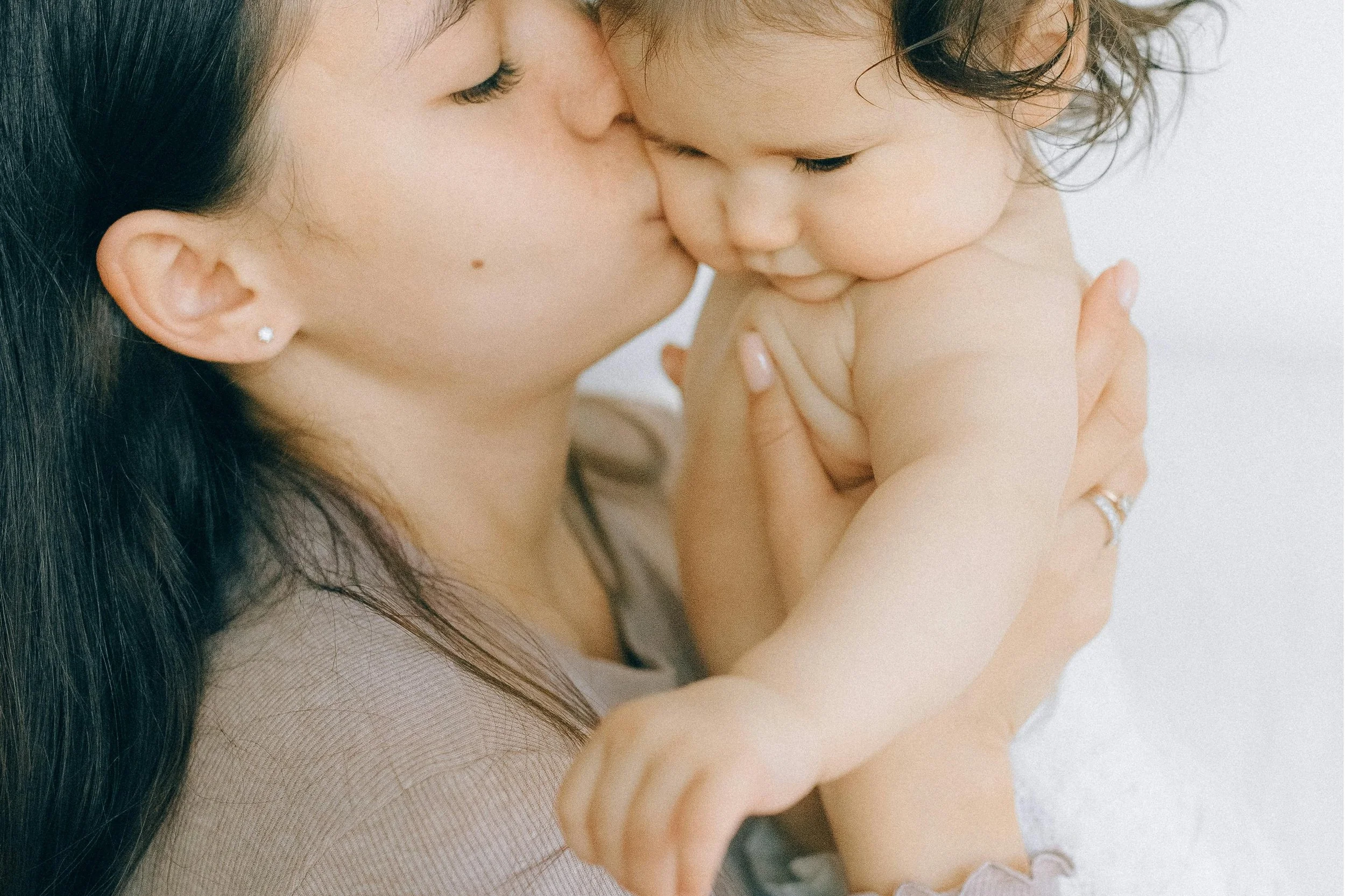 A woman with dark hair and earrings is holding a baby with curly hair close to her face, and they are touching noses in an intimate moment.