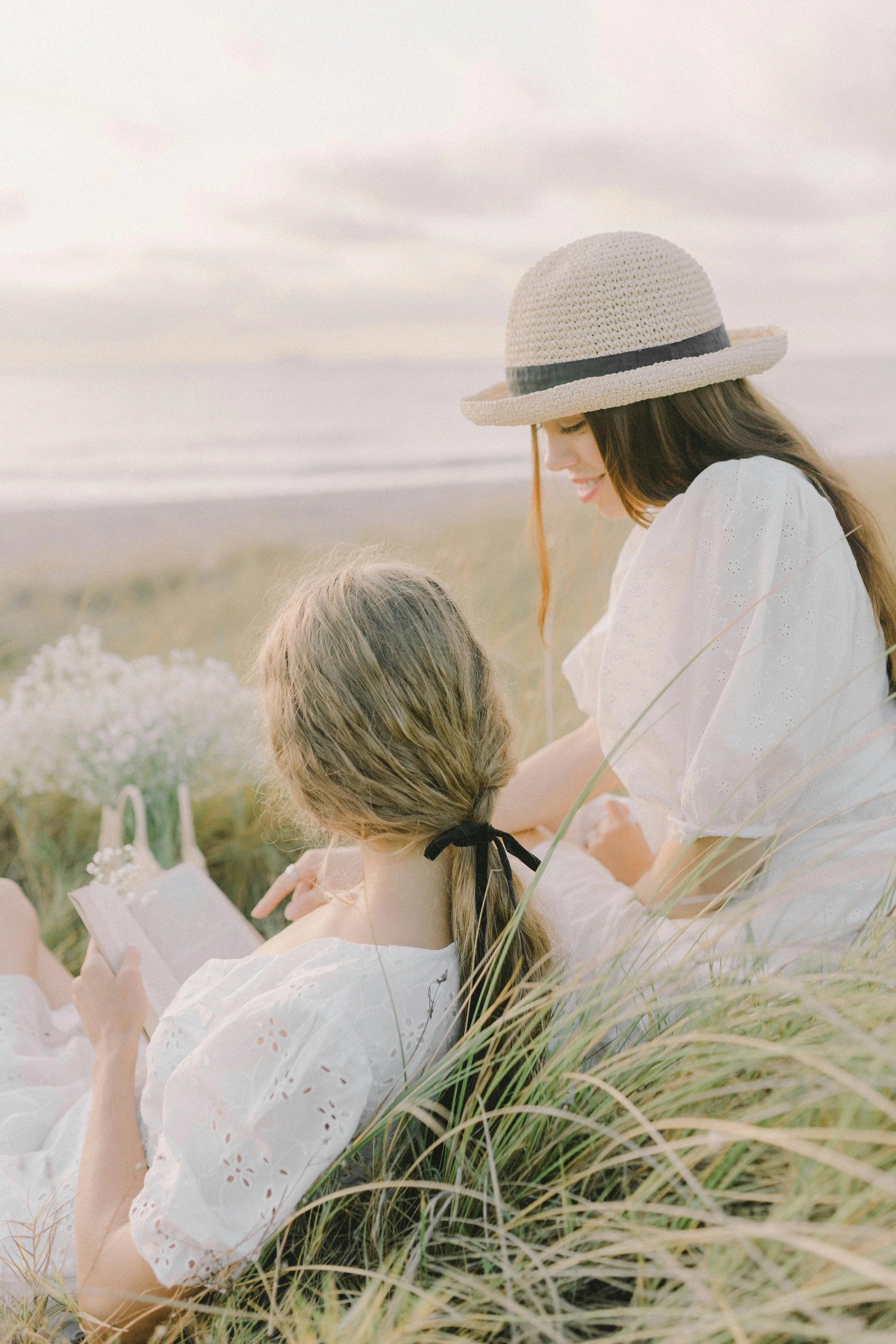Two women sitting in grass near the beach, one wearing a straw hat, smiling, and the other with a book.