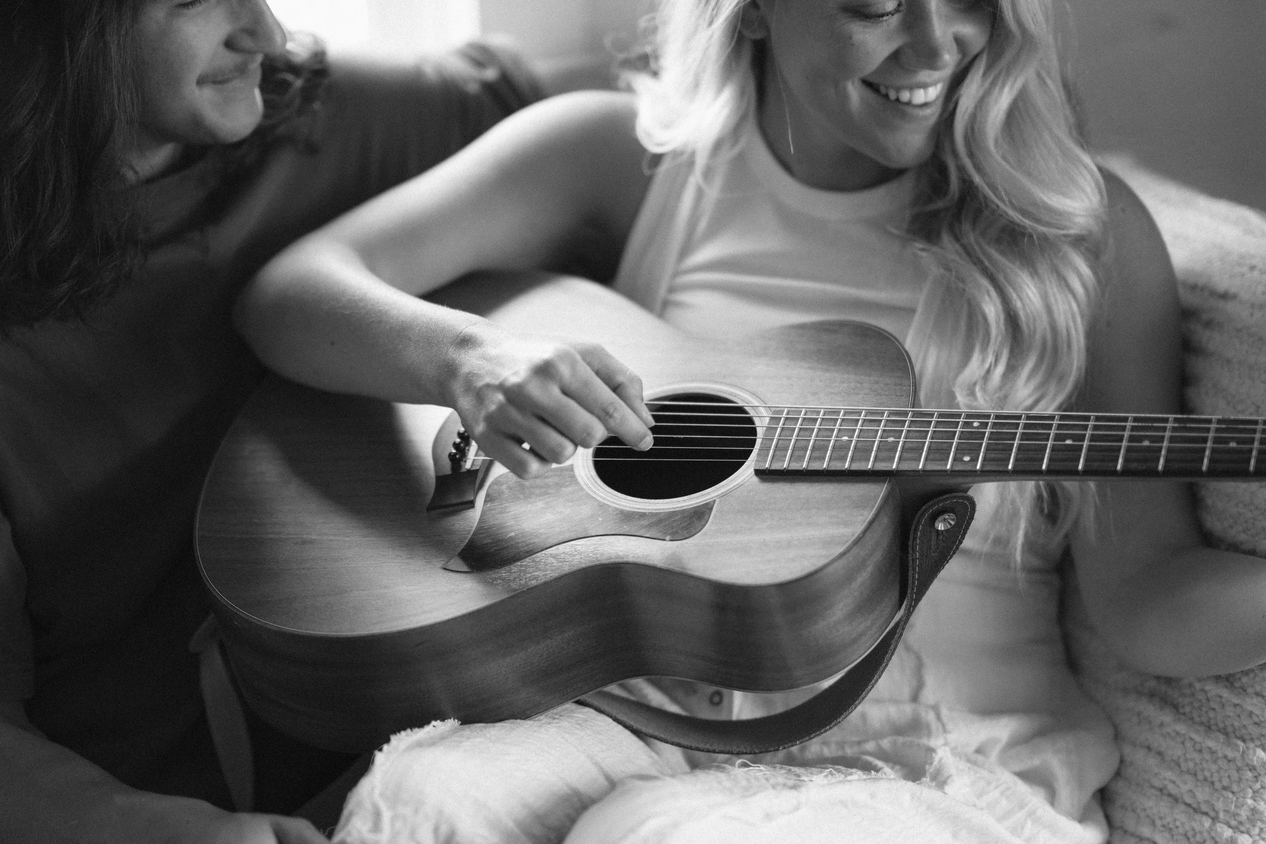 Two women sitting on a couch, one playing an acoustic guitar and the other smiling and watching.