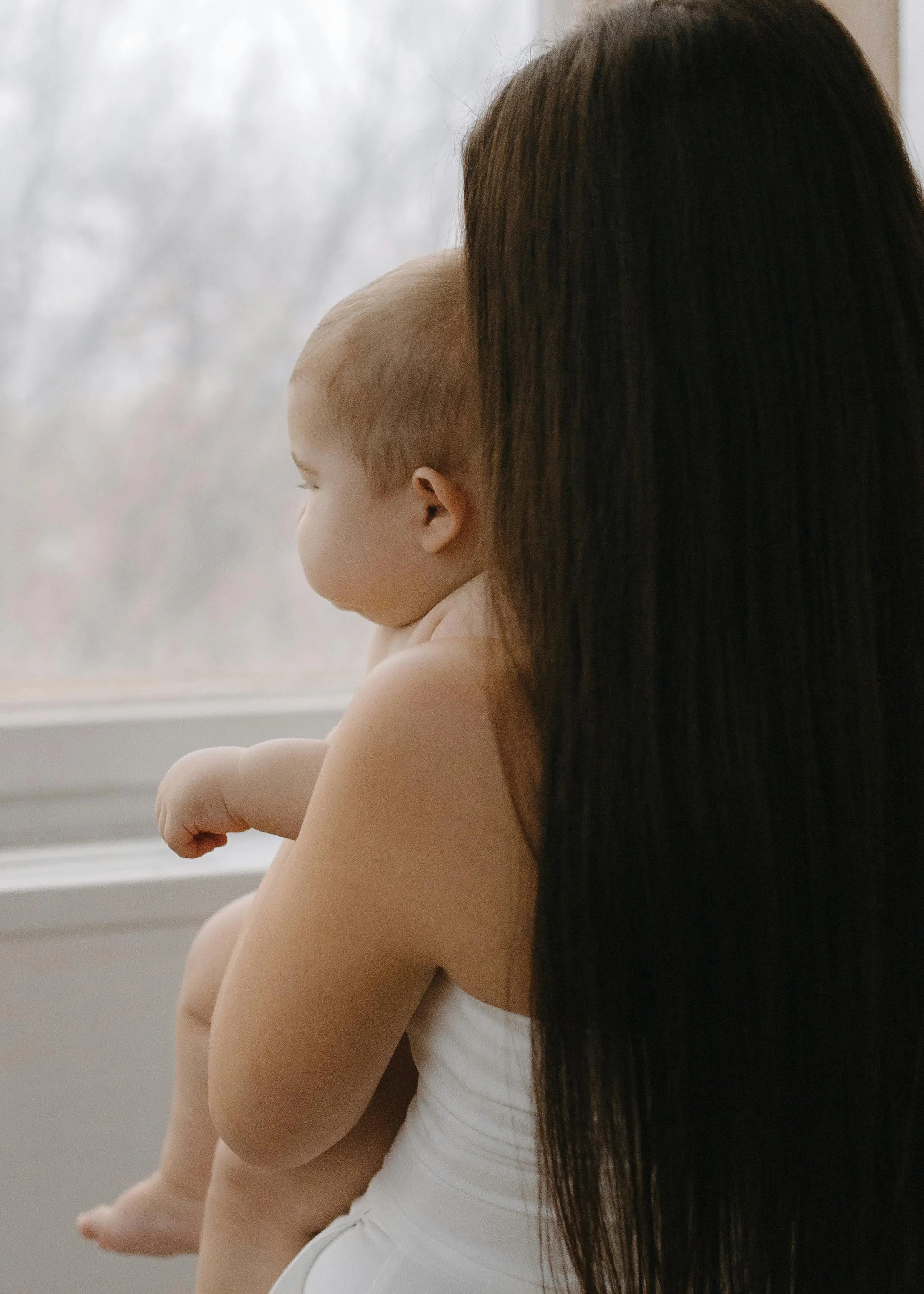 A woman holding a young child near a window, both facing outside, with the woman’s long dark hair and the child's light hair visible.