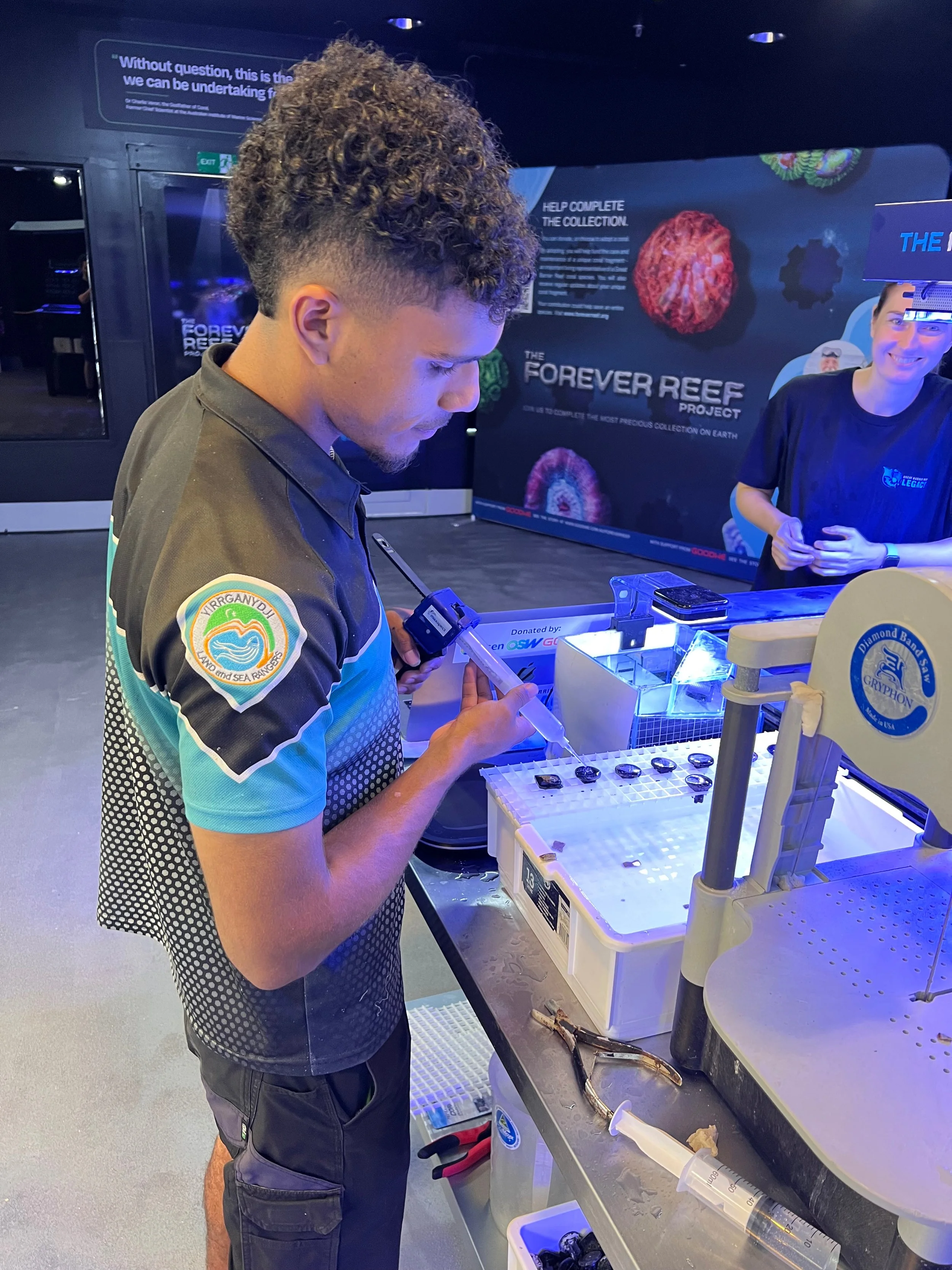 A young man with curly hair and a colorful shirt is working with scientific equipment at a display table, with a large poster behind him reading 'The Forever Reef Project.' Another individual is smiling in the background at an exhibit.