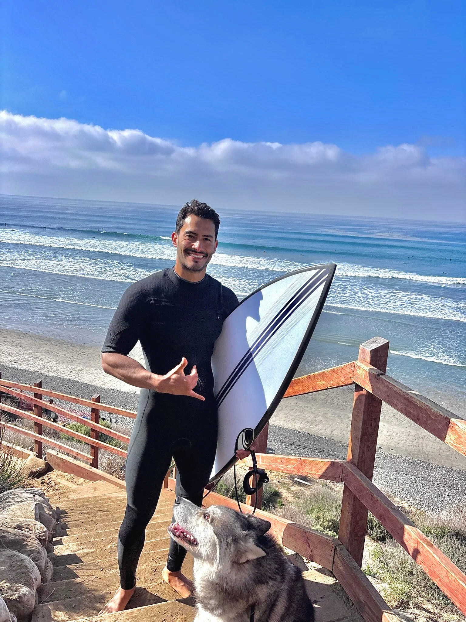 A smiling man holding a surfboard on a beach stairway, with a husky dog, ocean waves, and blue sky with clouds in the background.