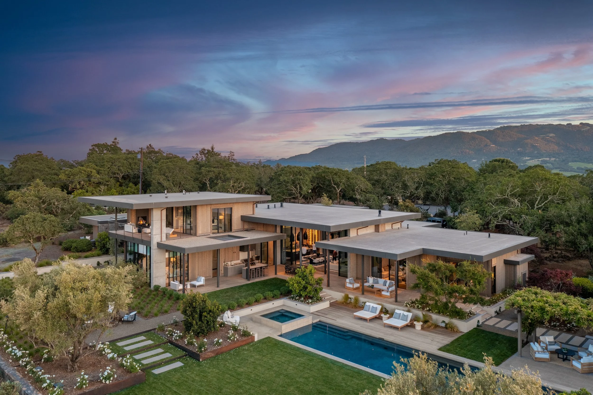 Modern house with large glass windows, outdoor pool, and landscaped yard at sunset, surrounded by trees and mountains in the background.