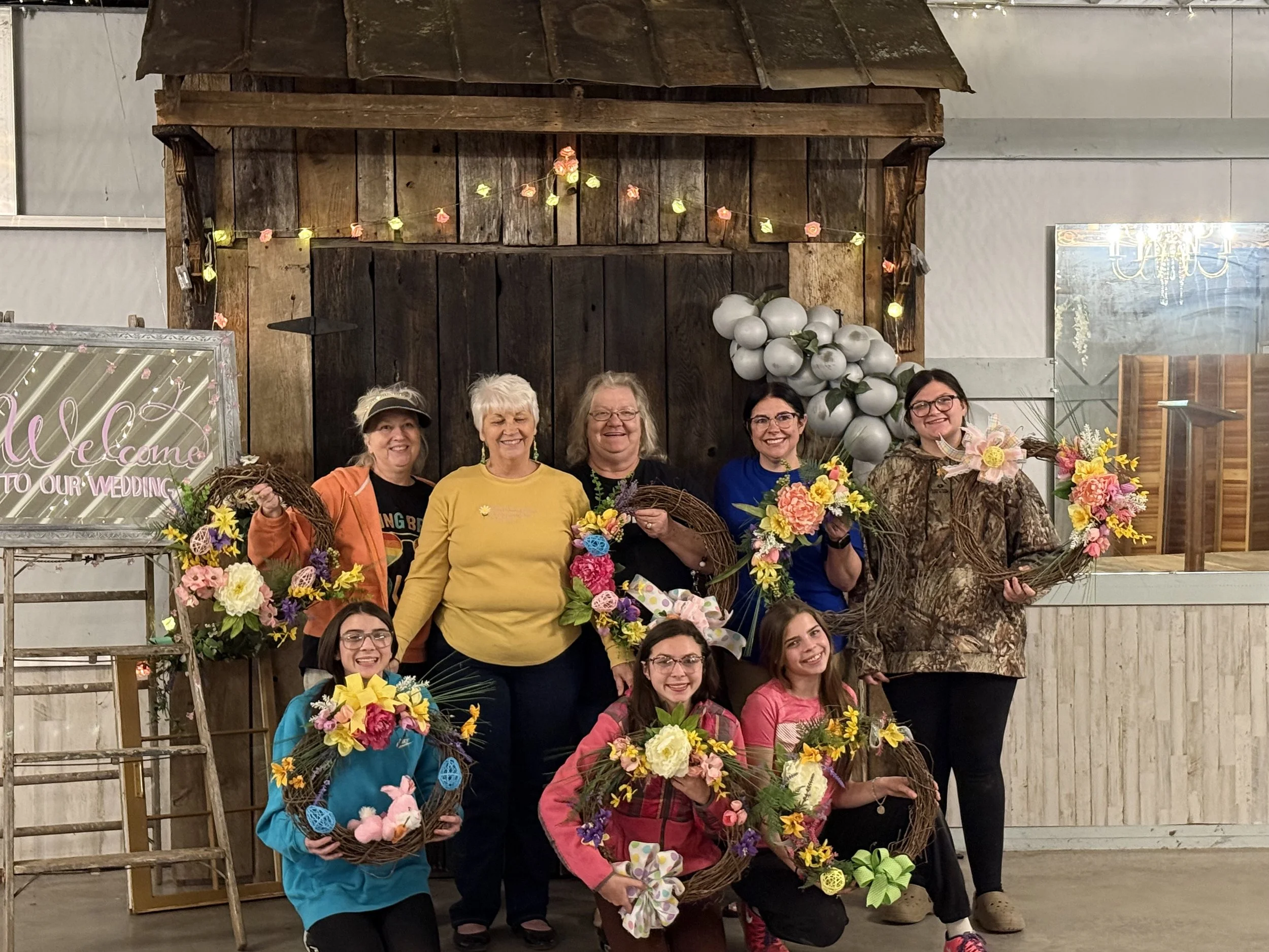 Group of women and girls at a wedding celebration holding floral wreaths and baskets, standing in front of a rustic backdrop with balloons and a sign that reads "Welcome to our wedding".