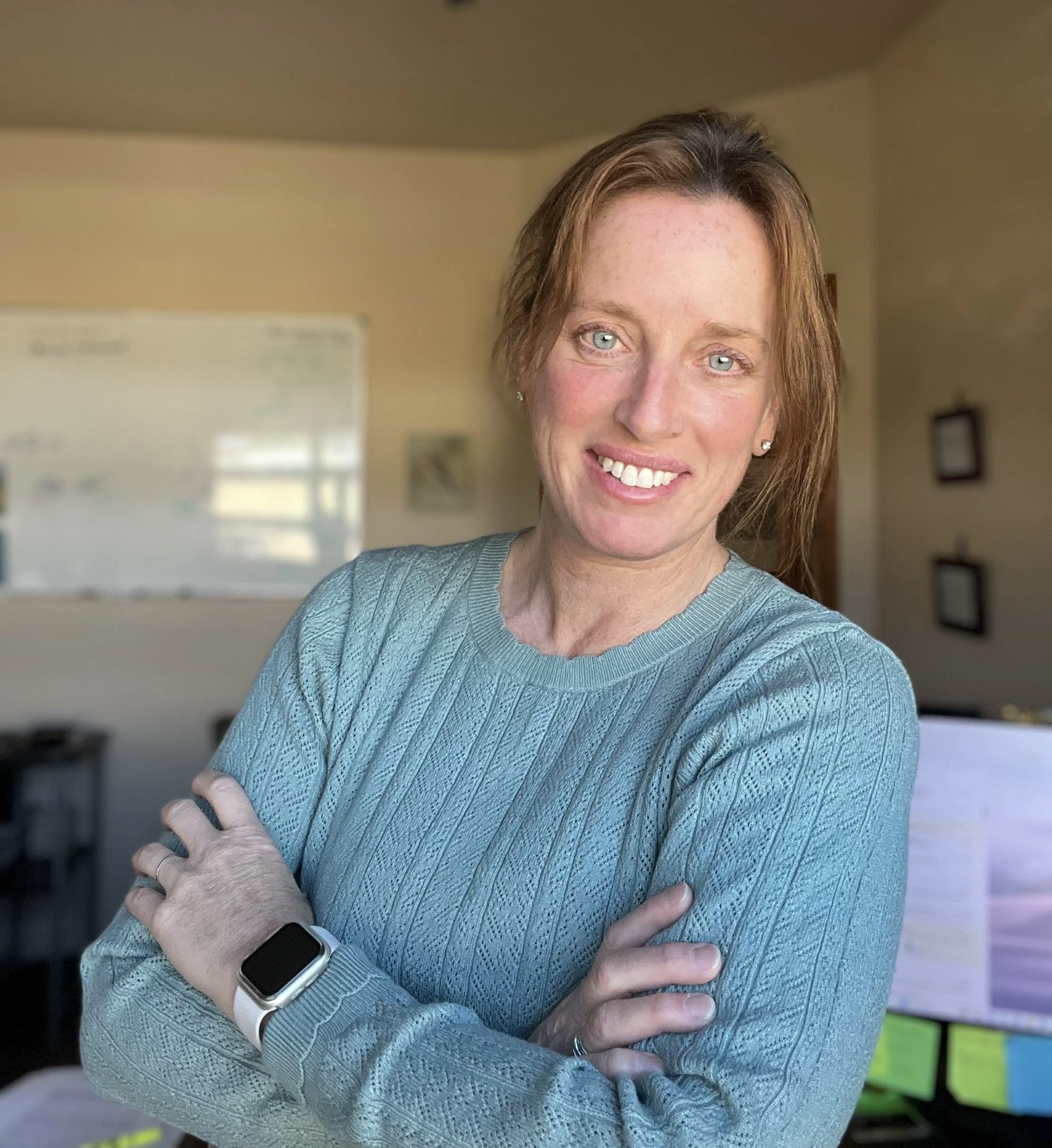 A woman with red hair and blue eyes smiling with arms crossed, wearing a gray sweater and a smartwatch, standing in a home office with a whiteboard and framed pictures in the background.