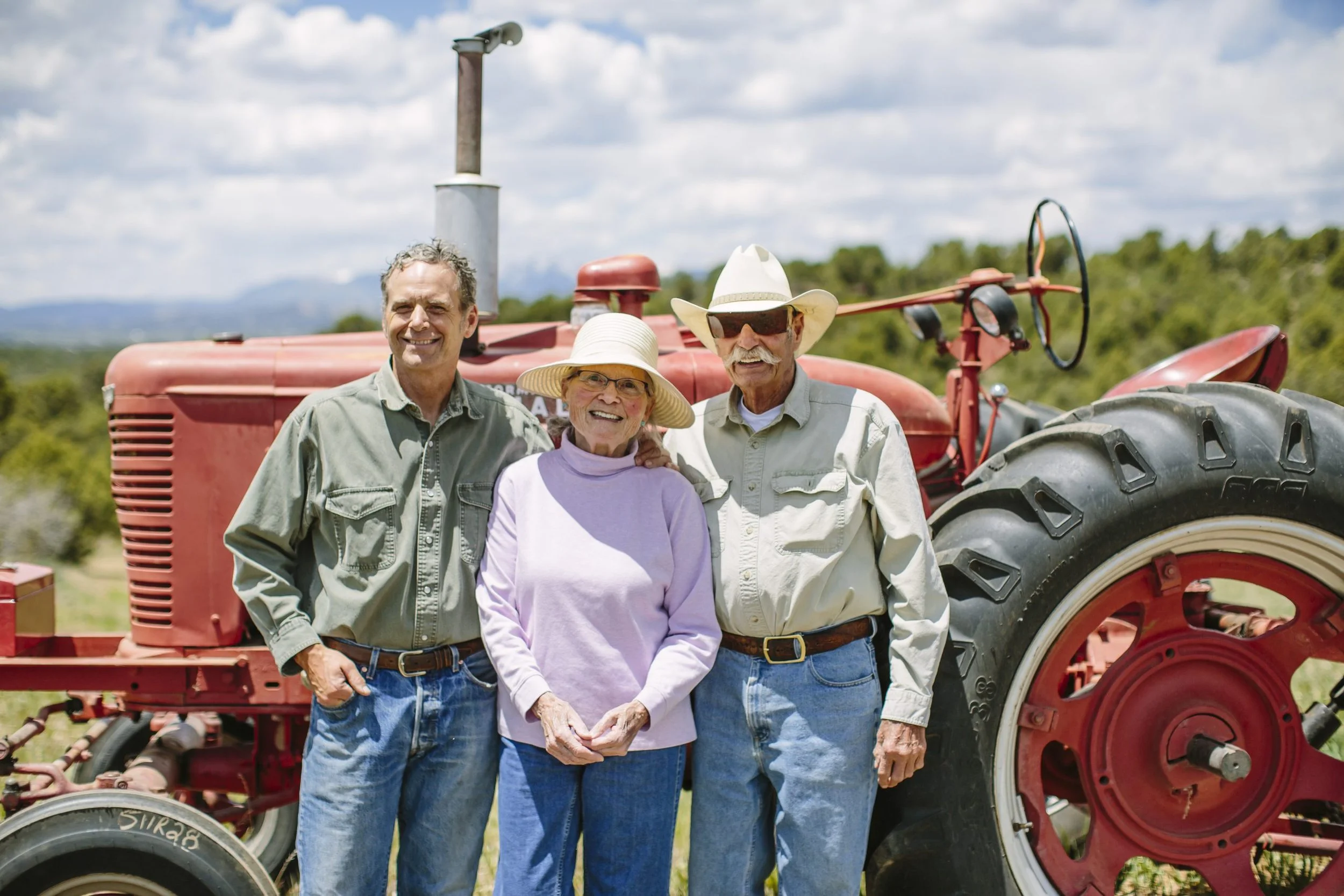 Three seniors, two men and one woman, standing together outdoors in front of a red tractor, smiling and posing for a photo on a sunny day with a partly cloudy sky and green landscape in the background.