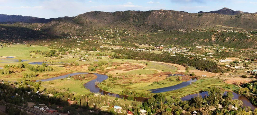 A winding river flowing through green fields and a small town with houses, set against rolling hills and mountains in the distance.