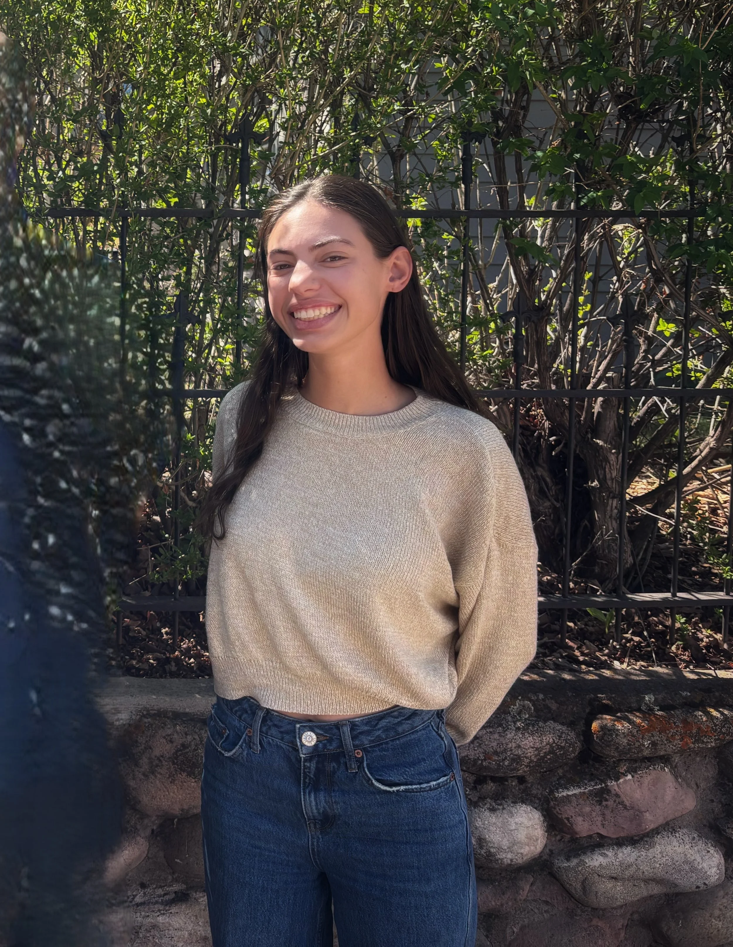 Smiling young woman wearing a navy Marmot puffer jacket standing outdoors with pine tree branches in the background.