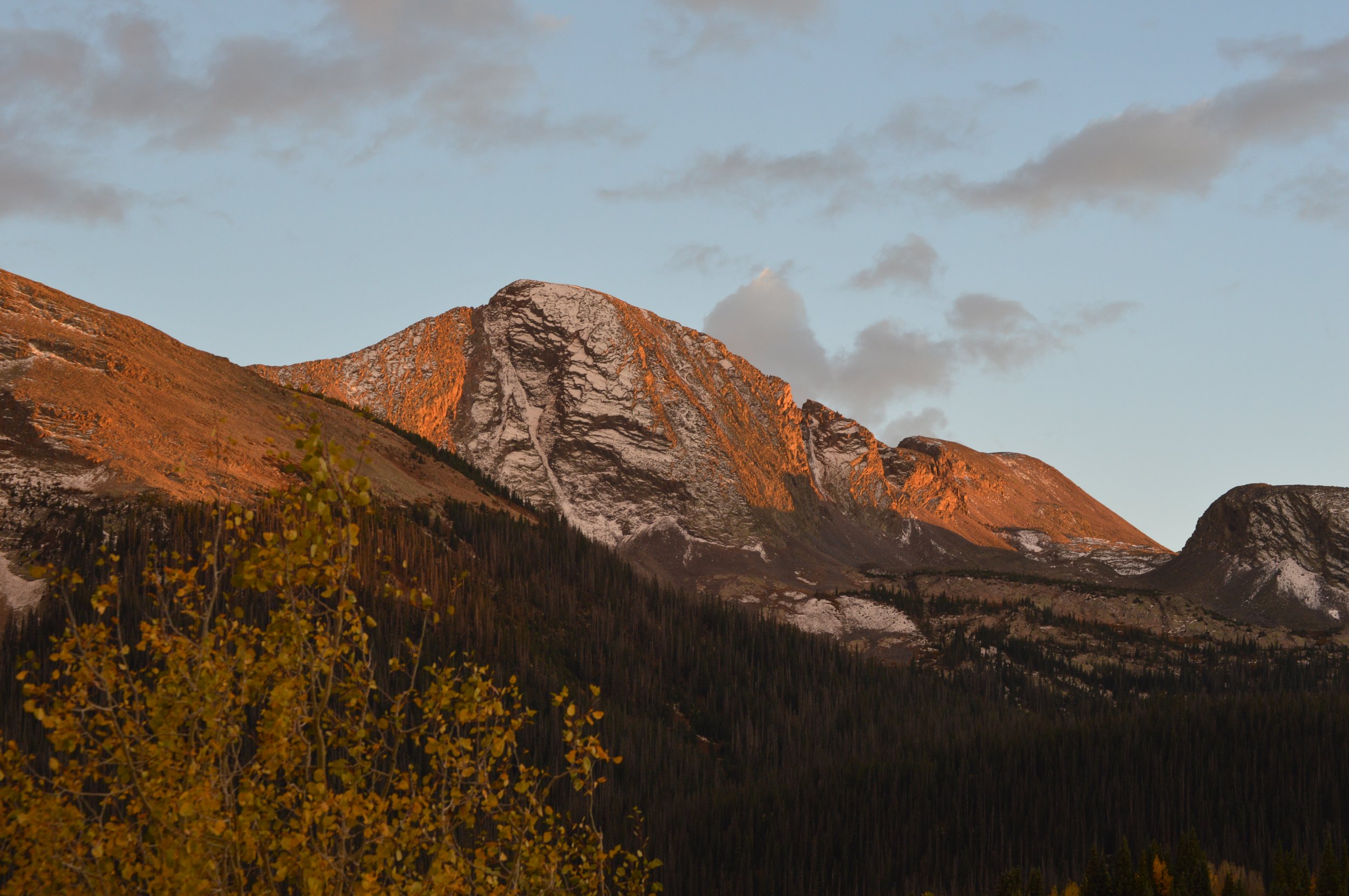 A mountain landscape during sunset with rocky mountain peaks illuminated by orange glow, surrounded by pine trees and yellow foliage in the foreground, and a partly cloudy sky above.
