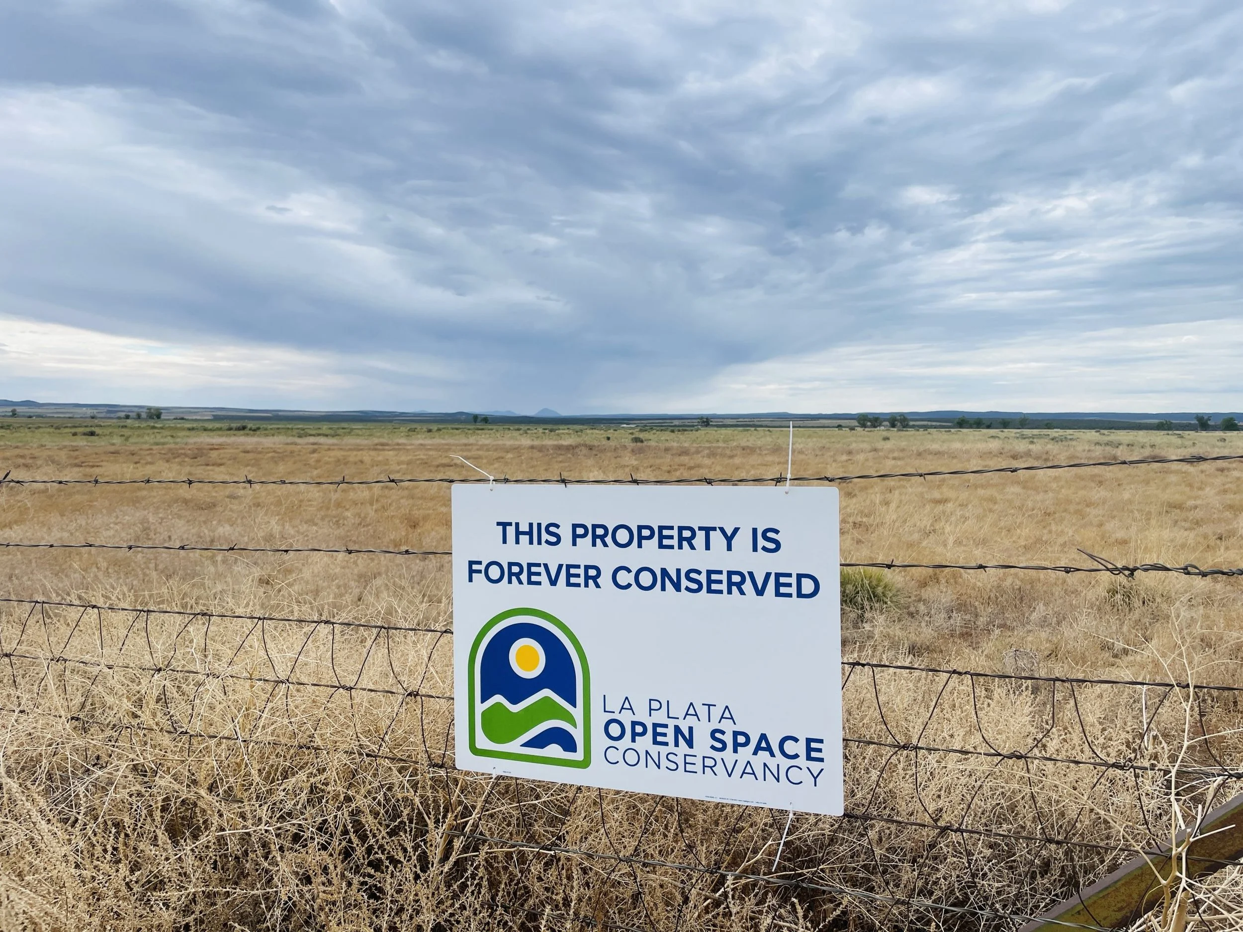 A sign on a wire fence in a dry field with cloudy sky in the background, reading 'This property is forever conserved' by La Plata Open Space Conservancy.