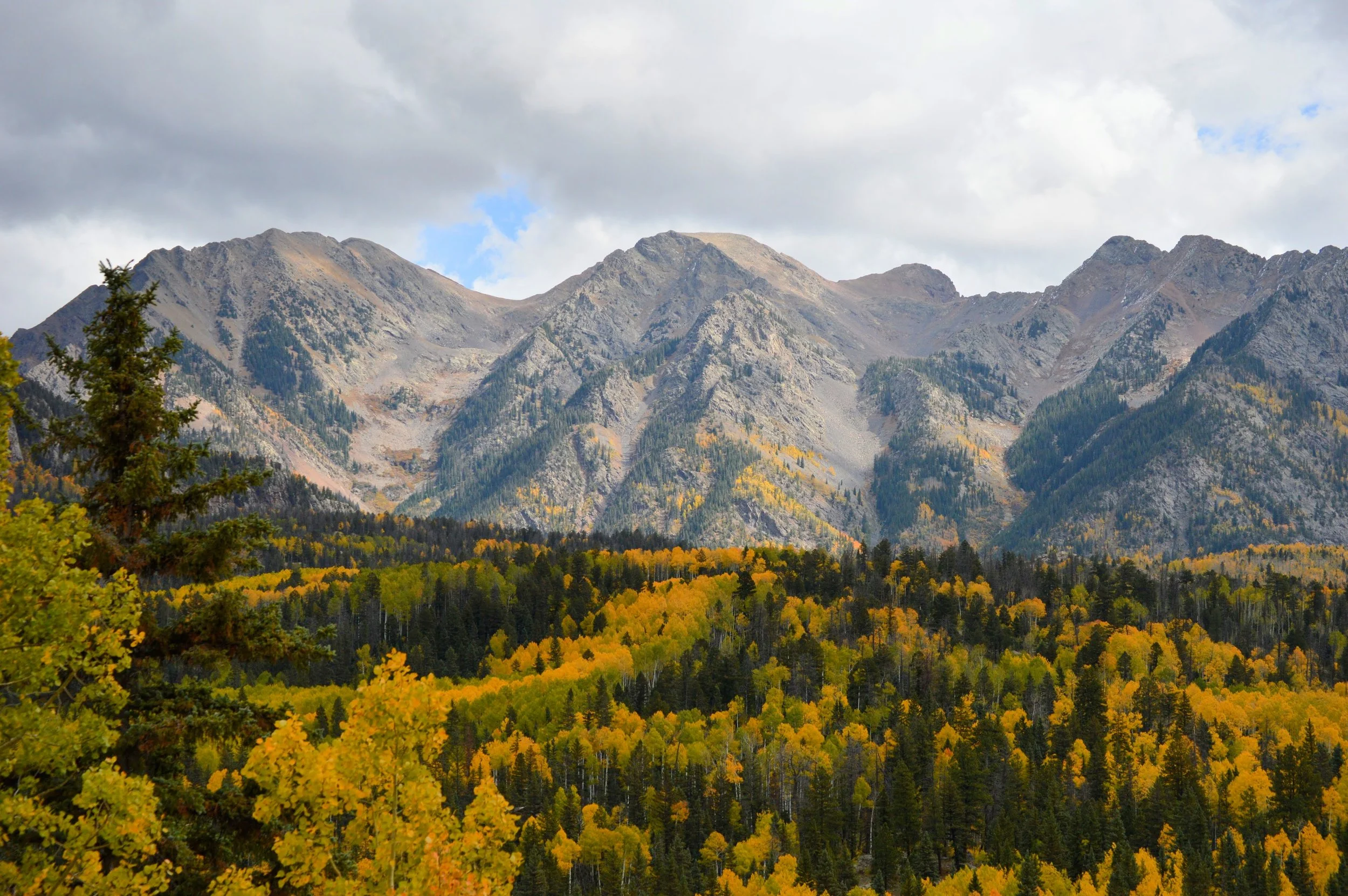 A mountain range with rocky peaks under a cloudy sky, surrounded by a forest with yellow, green, and some orange fall foliage.