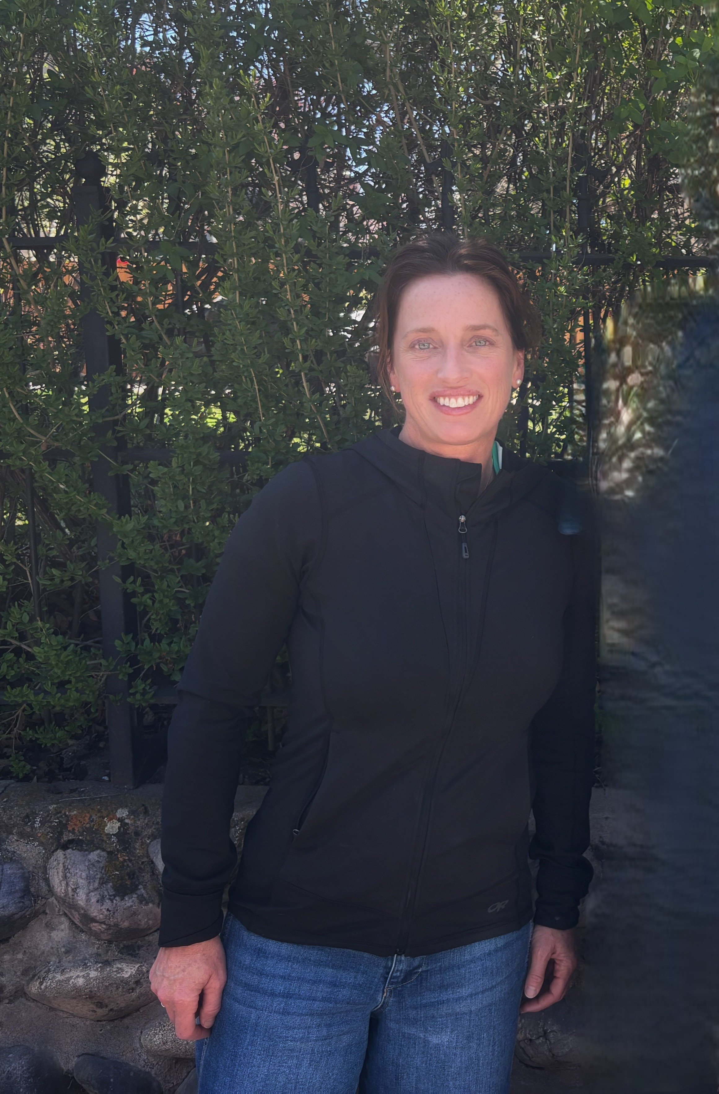 A woman with red hair and blue eyes smiling with arms crossed, wearing a gray sweater and a smartwatch, standing in a home office with a whiteboard and framed pictures in the background.