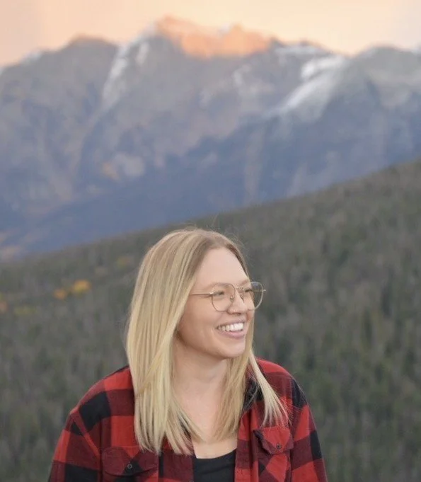 A smiling woman with blond hair and glasses standing outdoors with mountains in the background.