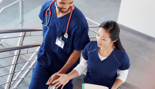 Two healthcare professionals, a male and a female nurse, walking down stairs in a hospital, engaging in conversation.