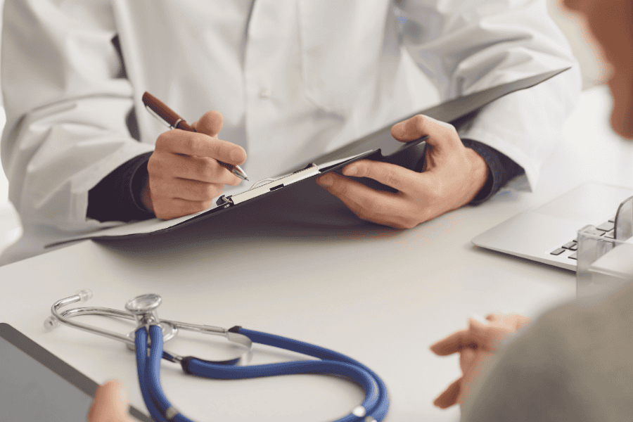 A healthcare professional in a white coat taking notes on a clipboard during a consultation with a patient. A stethoscope is on the table.