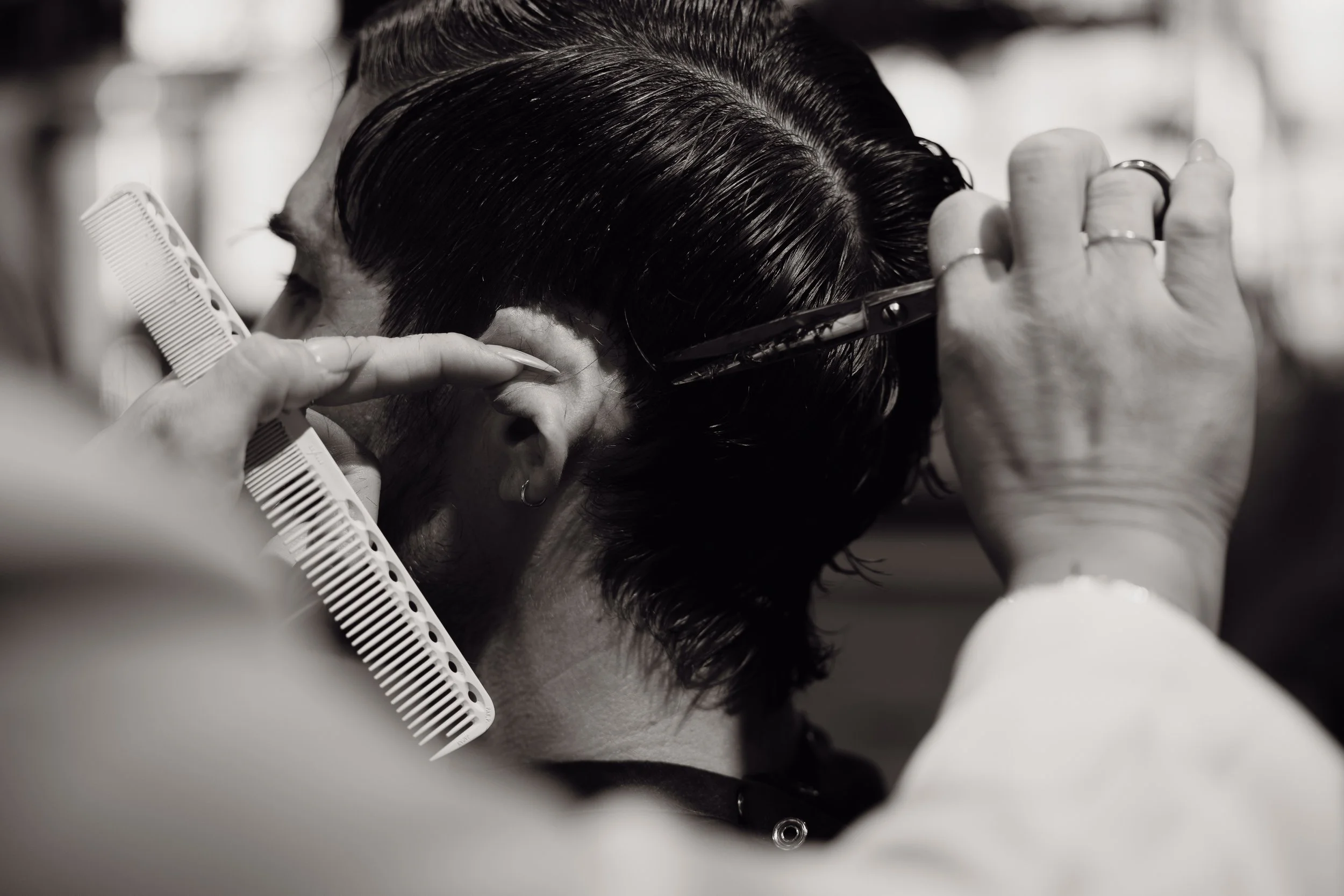 A person is cutting or styling another person's dark, short hair with scissors, comb, and hair clips in a salon or barbershop.
