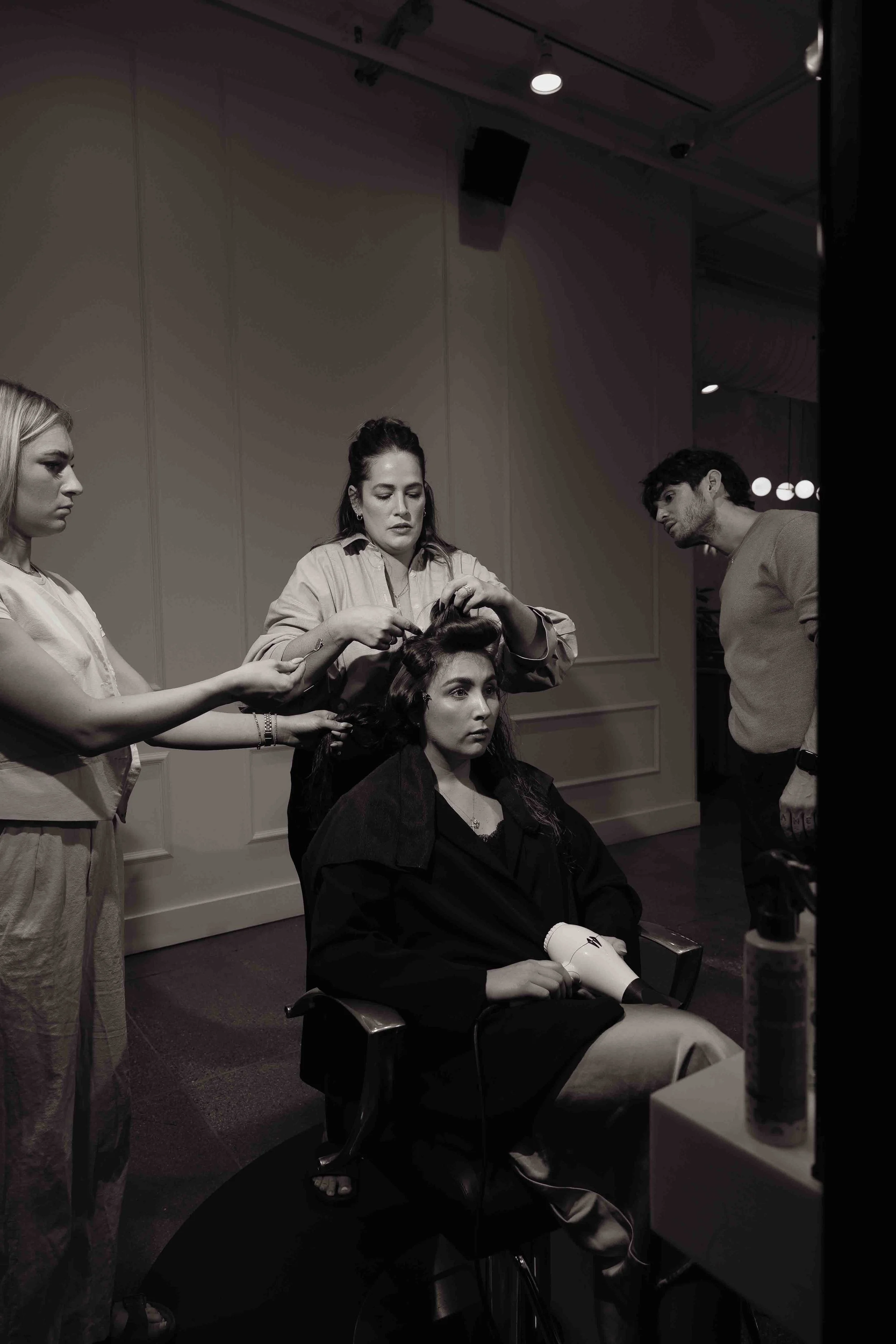 A woman is sitting in a salon chair with her hair being styled by two hairstylists, while a man observes. The scene appears to be in a salon or backstage setting, and the image is in black and white.