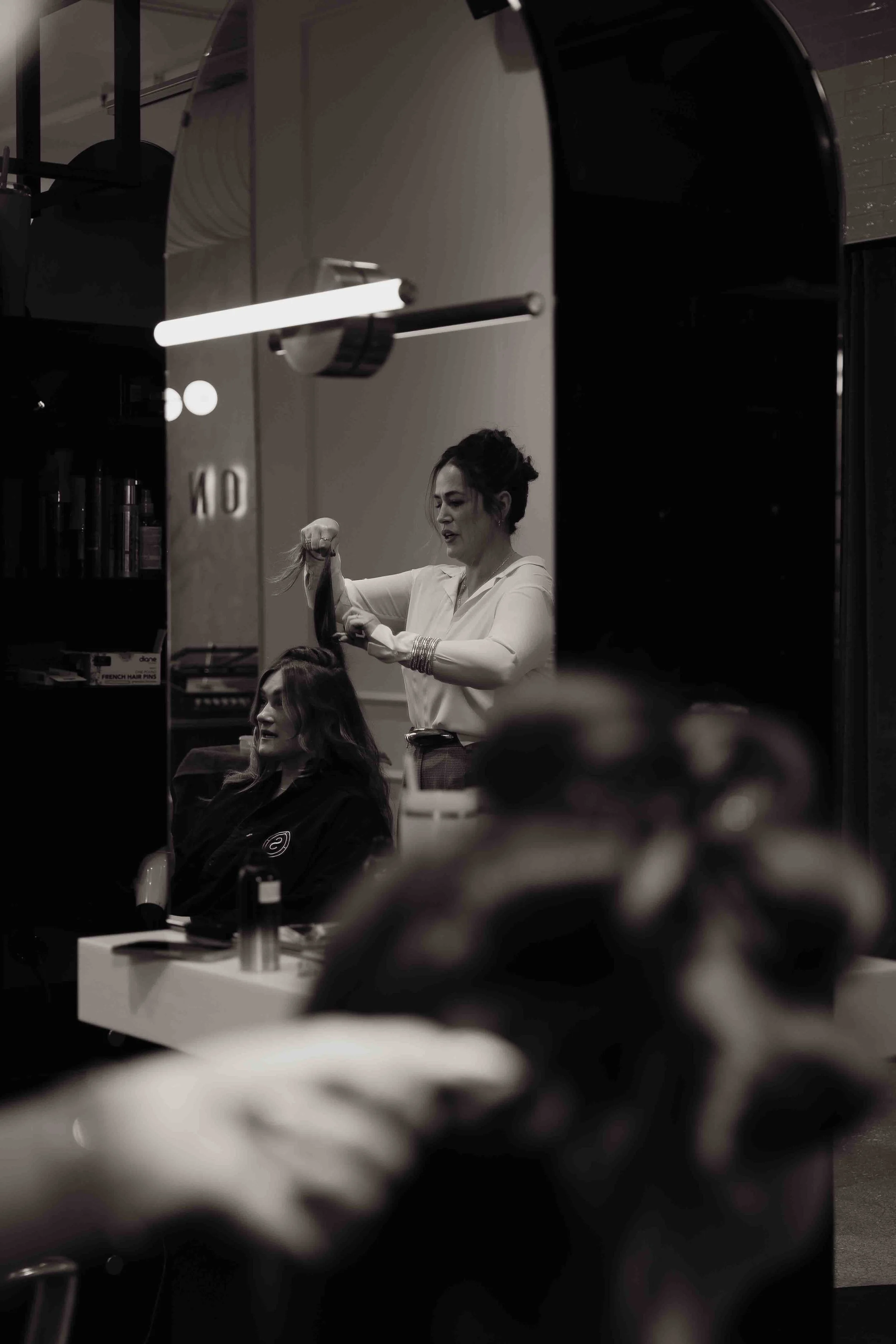A black and white photo of a woman getting her hair styled in a salon mirror.
