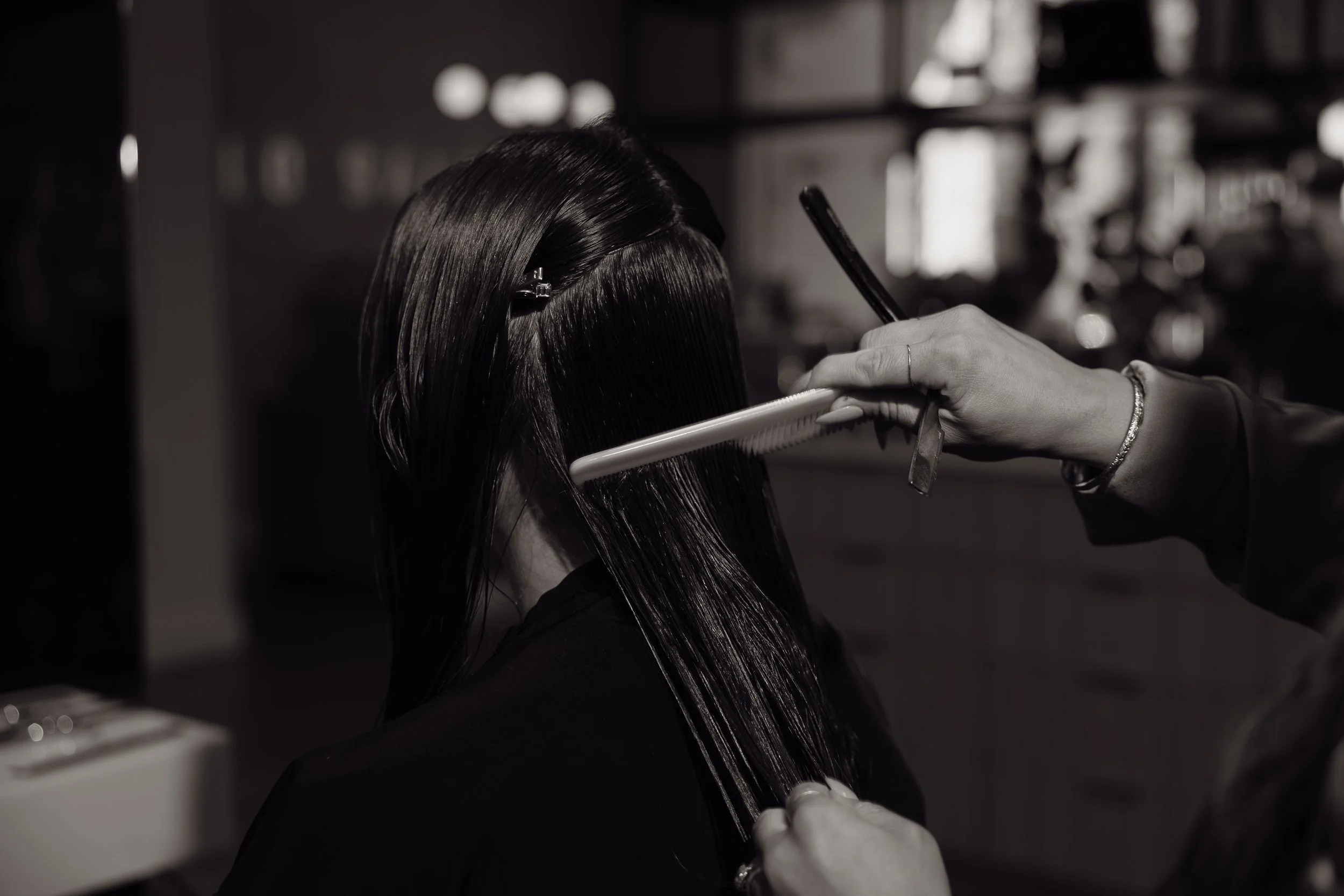 Black and white photo of a hairdresser straightening a woman's long, shiny hair with a flat iron and comb in a salon.
