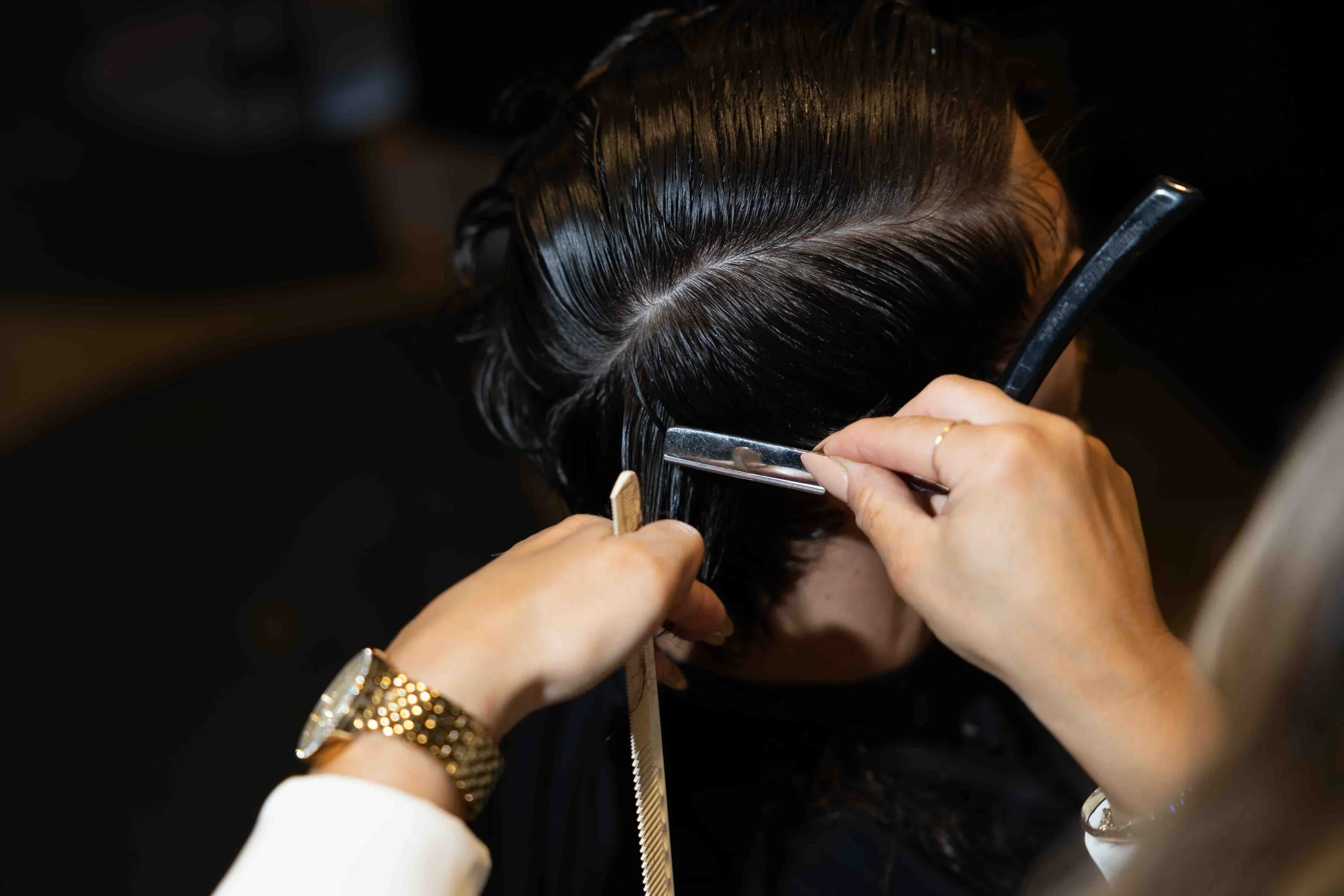 Hairdresser trimming and styling a client's dark, wet hair with scissors and a straight razor in a salon.