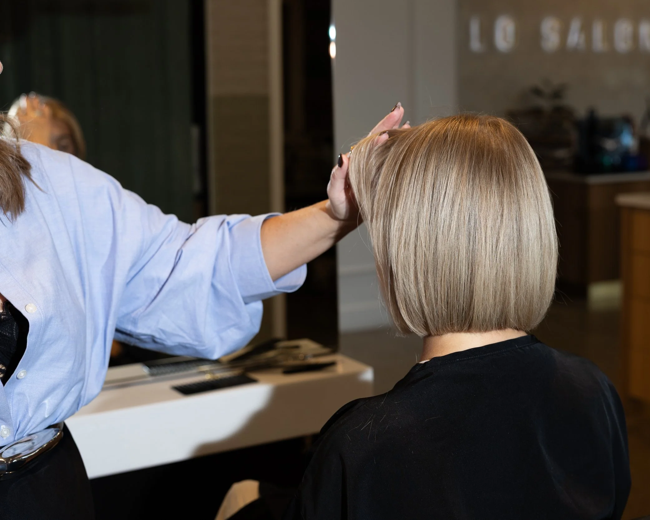 A woman in a black shirt is having blonde hair cut or styled by a hairstylist in a salon. The hairstylist, wearing a light blue shirt, is touching the woman's hair.