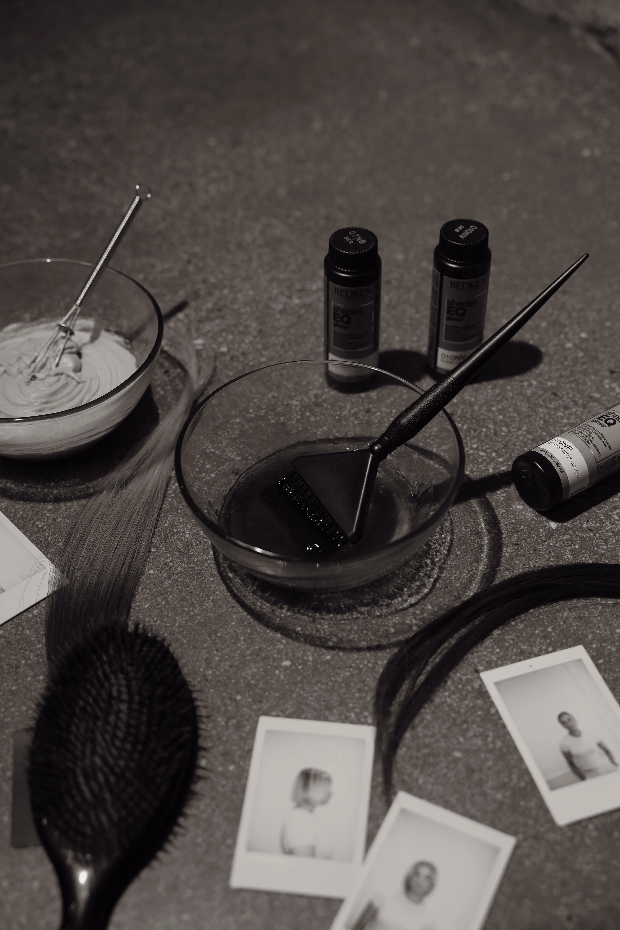 Hair coloring supplies including bowls with dye, brushes, hair clips, color bottles, and black-and-white photographs on a countertop.