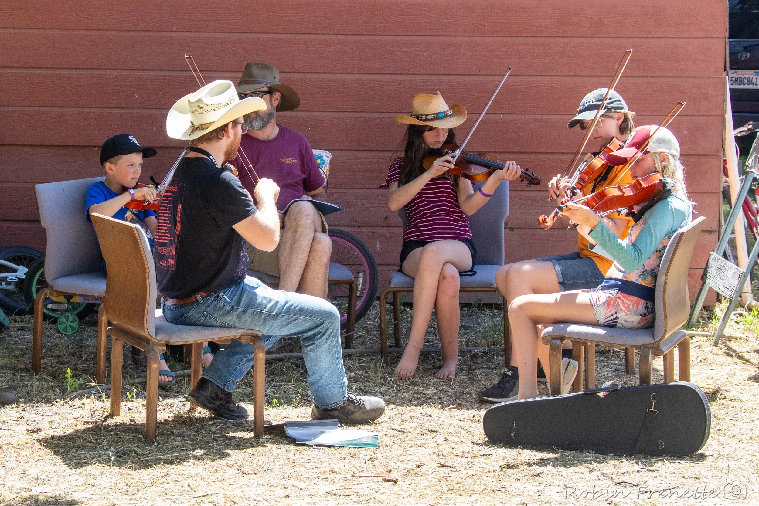 Kids sitting outdoors, playing string instruments, and singing, at the kids on bluegrass workshop at the Parkfield Bluegrass Festival