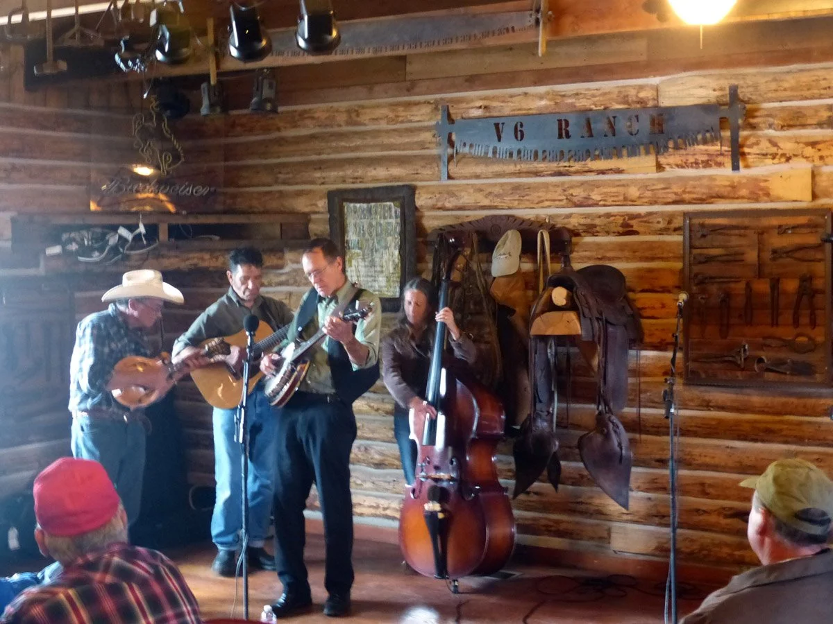 Four musicians performing on a wooden stage in the rustic Parkfield Cafe, playing guitars, a double bass, and a mandolin, with an audience seated in front during the Parkfield Bluegrass Festival