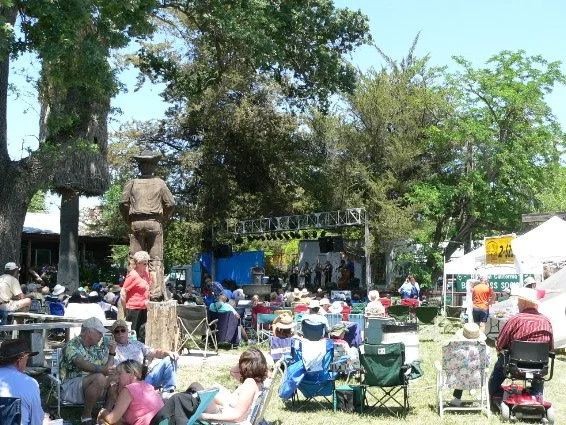 Old photo of people attending the Parkfield Bluegrass Festival, sitting on chairs and blankets, watching a stage with musicians, under trees, with tents.