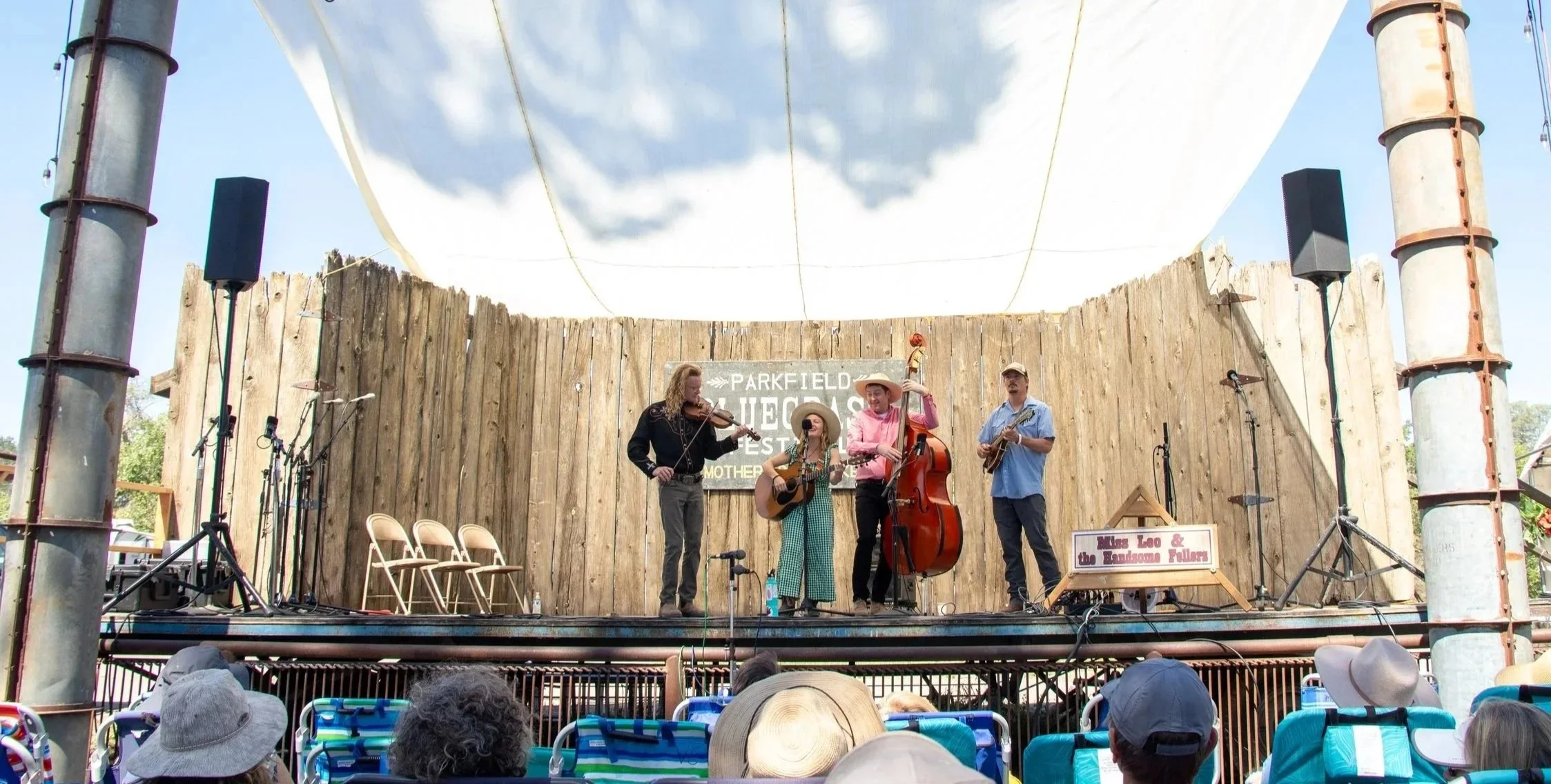 Miss Leo and the Handsome Fellers performing at the Parkfield Bluegrass Festival. The band includes a violinist, a guitarist, a bassist, and a mandolin player. The audience is seated in front, wearing hats and sunglasses, watching the performance.