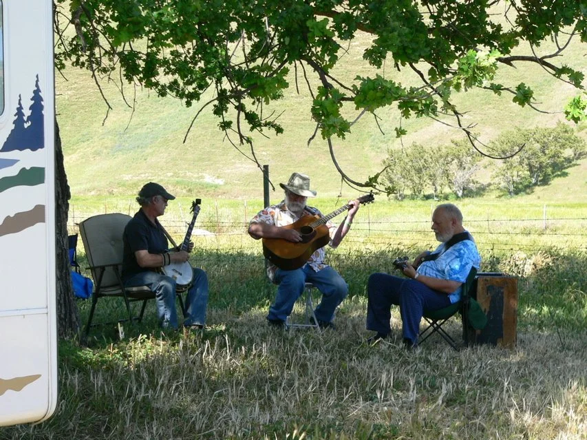 Three men sitting outdoors under a tree, playing guitars in the Parkfield Bluegrass Festival campsire