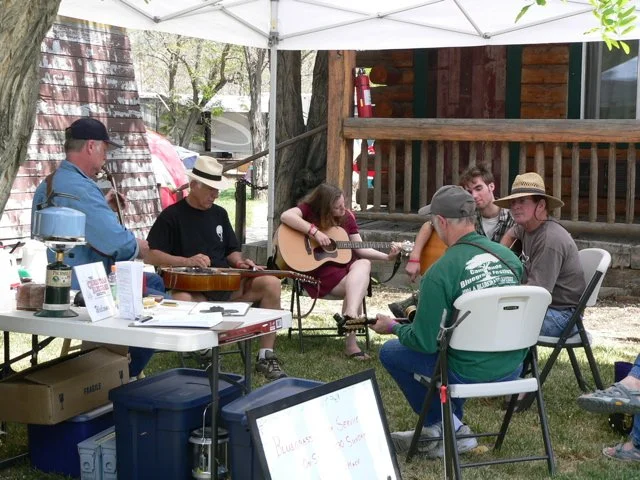 Group of people sitting under a white canopy outdoors, playing and listening to guitars, in front of the Parkfield Lodge during the Parkfield Bluegrass Festival