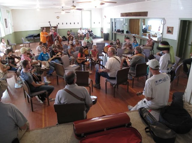 Group of people, including children and adults, gathered in a circle in the historic Parkfield Community Hall, some playing guitars, singing, or listening, with musical instruments during the Parkfield Bluegrass Festival