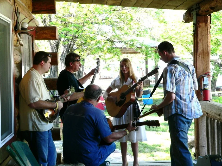 A group of five people playing musical instruments on the Parkfield Lodge porch with green trees and sunlight in the background during the Parkfield Bluegrass Festival