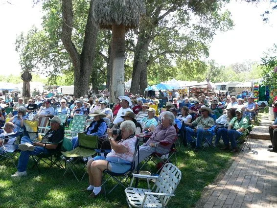 Old photo of the audience at the Parkfield Bluegrass Festival seated outdoors on lawn chairs under trees, with tents and booths in the background.