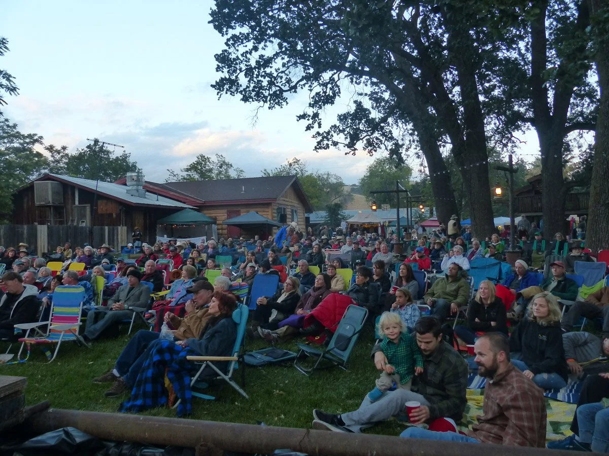 People gathered outdoors during the Parkfield Bluegrass Festival, sitting on chairs and blankets under trees, with the Parkfield Cafe and string lights in the background.