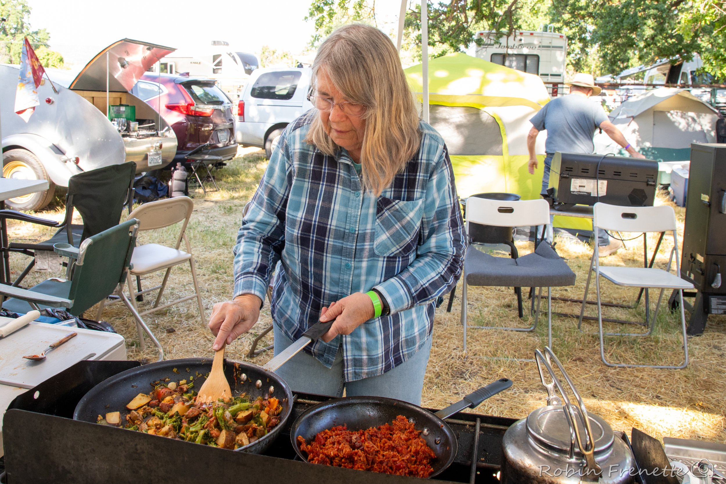 A woman in a plaid shirt cooking vegetables and rice in a frying pan outdoors, with chairs, tents, and vehicles in the background at the Parkfield Bluegrass Festival campsite