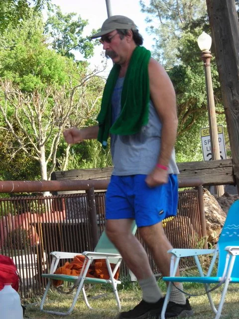 Dancin' John dancing at the Parkfield Bluegrass Festival, with a green towel draped around his neck. He is wearing sunglasses and walking past a row of outdoor chairs and a fence, with trees and a lamppost in the background.