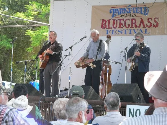 Old photo of three musicians performing on stage at the Parkfield Bluegrass Festival, with a banner and trees in the background, audience watching