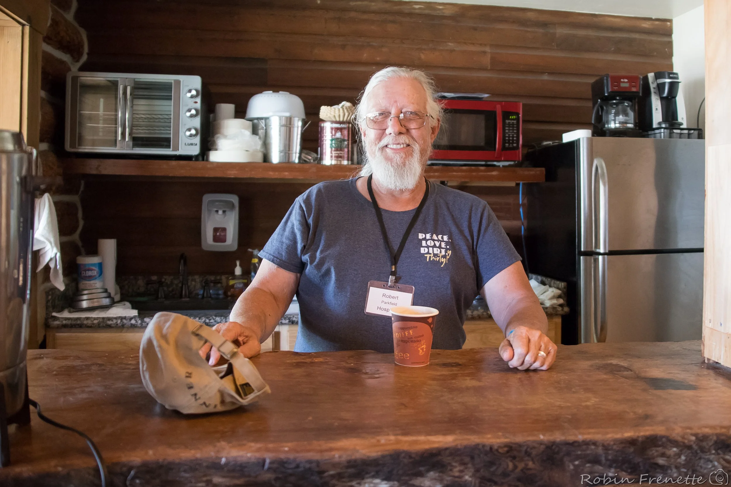 A smiling Parkfield Bluegrass Festival Volunteer, wearing glasses and a blue T-shirt, standing behind a wooden counter in a rustic kitchen, holding a paper cup of coffee and a beige bag.
