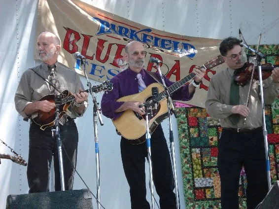 Old photo of the Roland White band performing on stage at the Parkfield Bluegrass Festival, with one playing a mandolin, another playing an acoustic guitar, and the third playing a violin