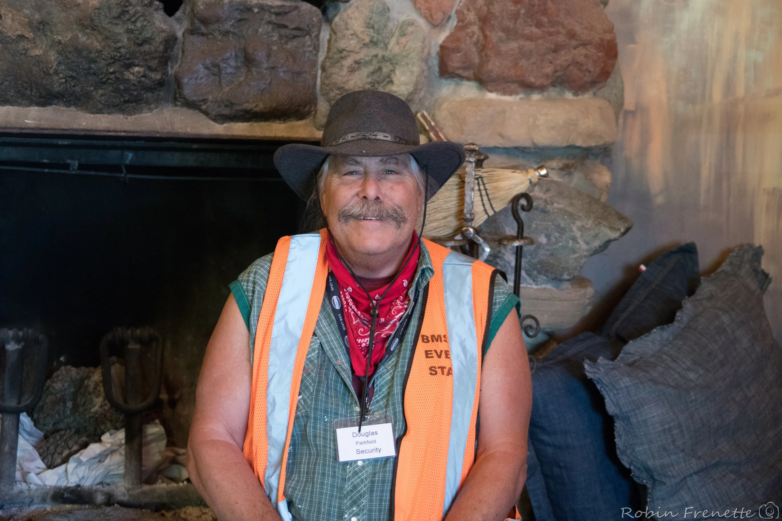 A Parkfield Bluegrass Festival volunteer in a cowboy hat, a green plaid shirt, an orange safety vest, a red bandana, and a security badge, sitting indoors in front of a stone fireplace with pillows and decorative items nearby.
