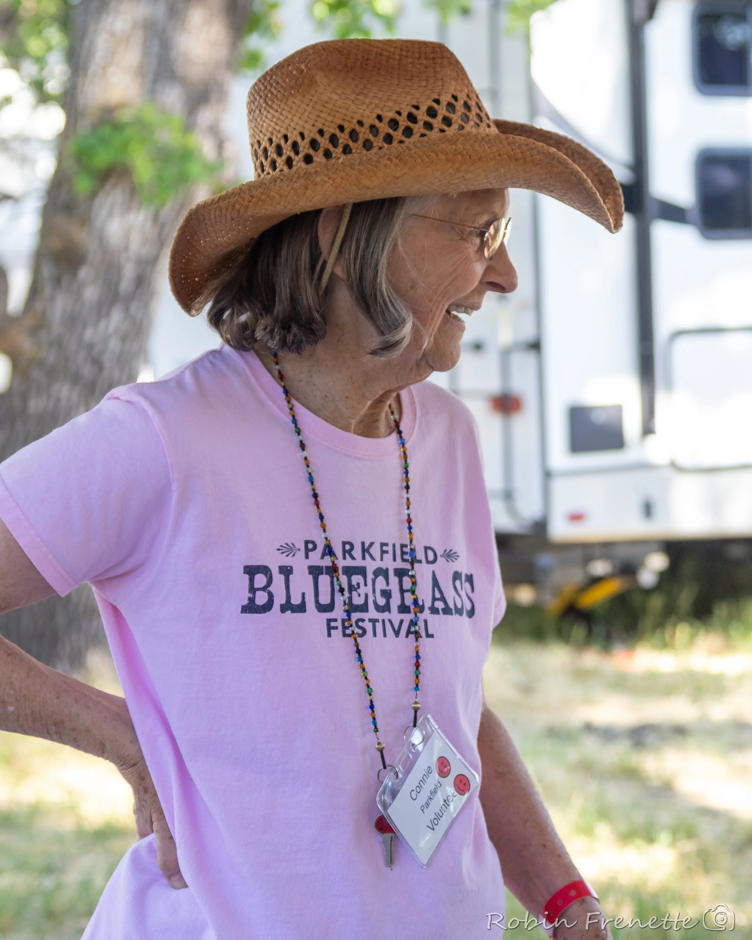 Connie Moxness wearing a straw cowboy hat, glasses, a pink t-shirt with 'Parkfield Bluegrass Festival' printed on it, and a multicolored beaded necklace with a volunteer ID badge hanging from neck, at the Parkfield Bluegrass Festival.