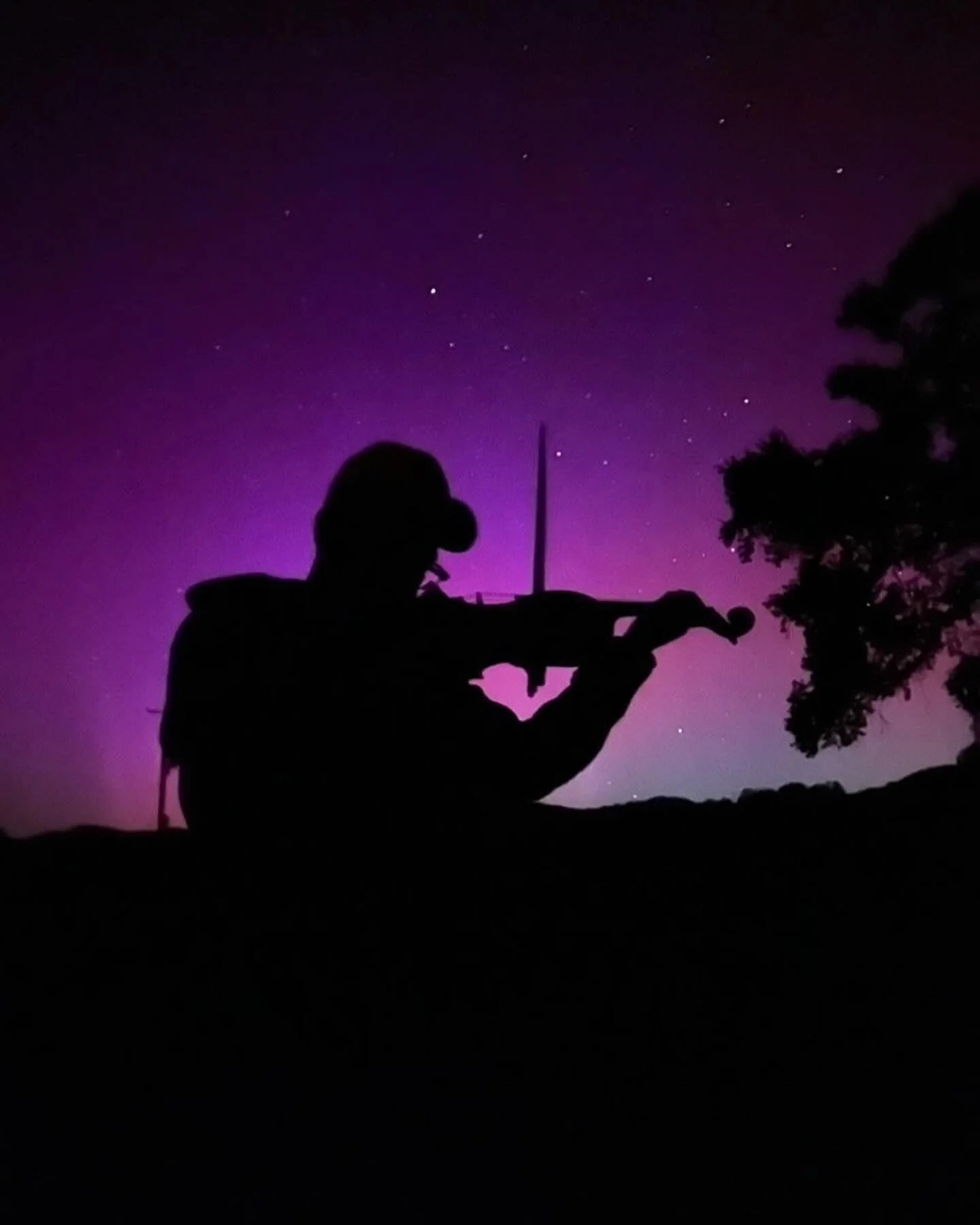 Silhouette of a person playing a fiddle outdoors at night with the aurora borealis in the background